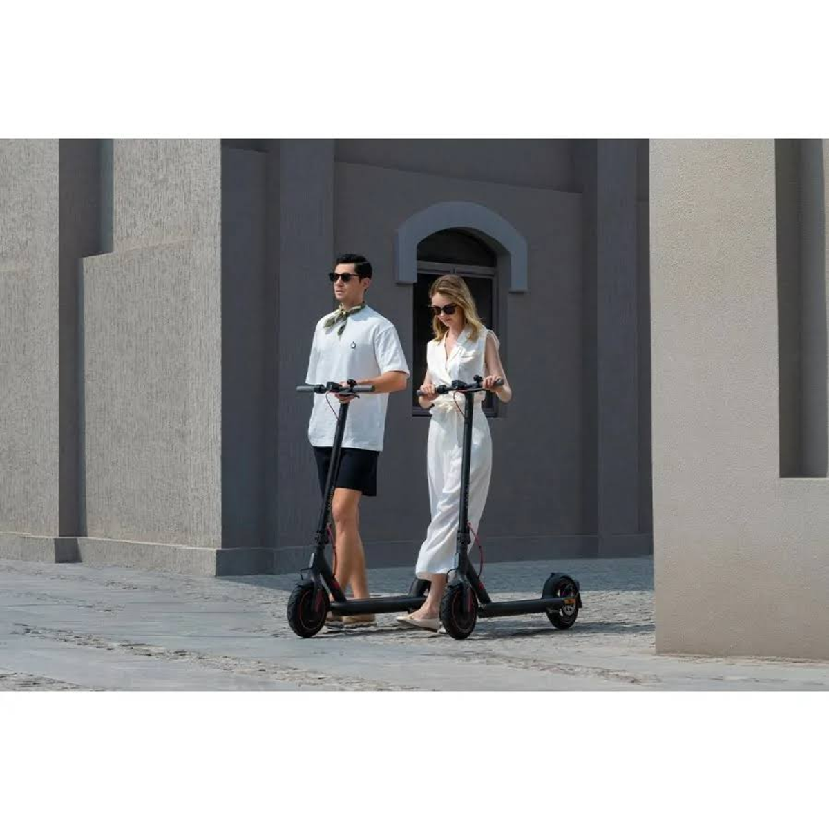 Un hombre y una mujer montan scooters eléctricos en un camino pavimentado. Edificios en el fondo.