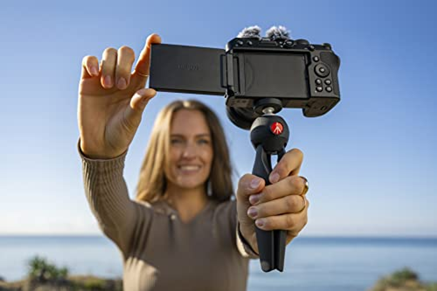 Eine Frau hält eine Kamera mit Mikrofon an einem Stativ, mit blauem Himmel im Hintergrund.