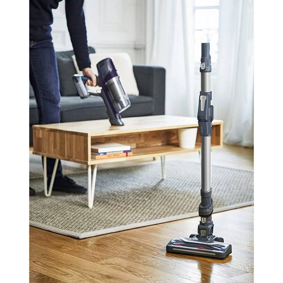Eine Person, die einen kabellosen Staubsauger benutzt. Ein Holztisch und ein grauer Teppich sind ebenfalls im Bild.
