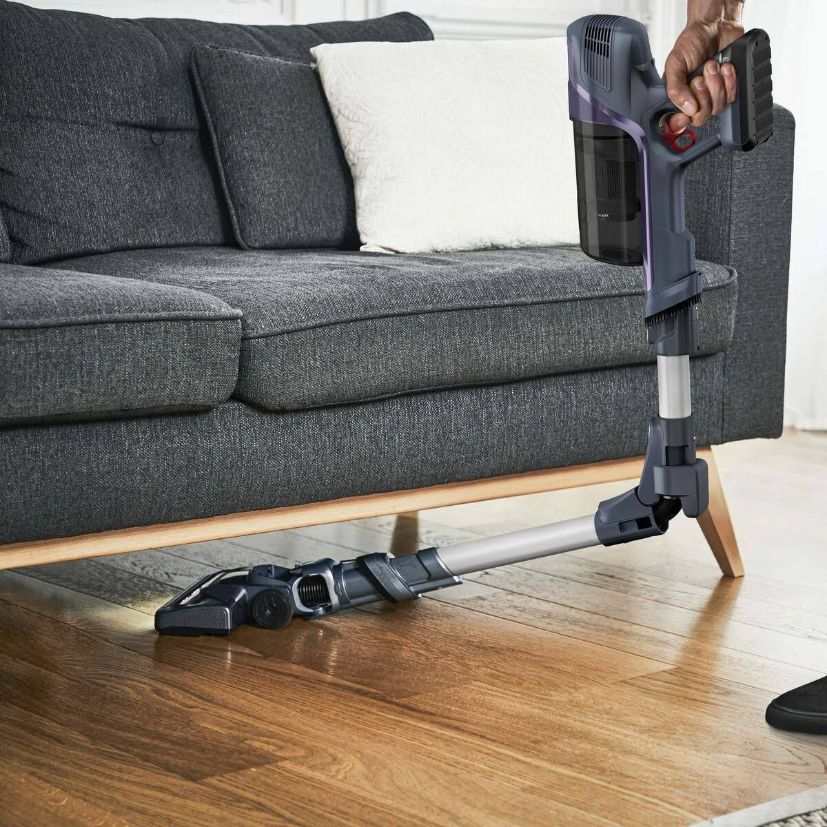 Una persona che utilizza un aspirapolvere con una testa angolata su un pavimento in legno, divano grigio dietro.