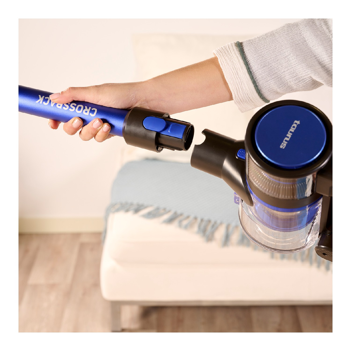 Una mano conectando un tubo azul de aspiradora al cuerpo principal. Un sofá en el fondo.