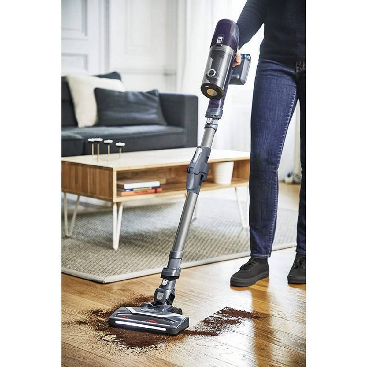 Una persona usa un aspirapolvere senza fili su un pavimento in legno con detriti.