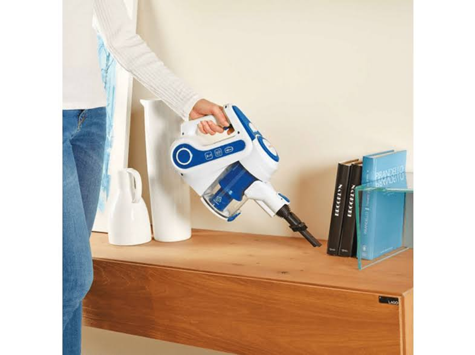 Una persona usa un aspirapolvere portatile blu e bianco su una superficie di legno, vicino a libri e un vaso.