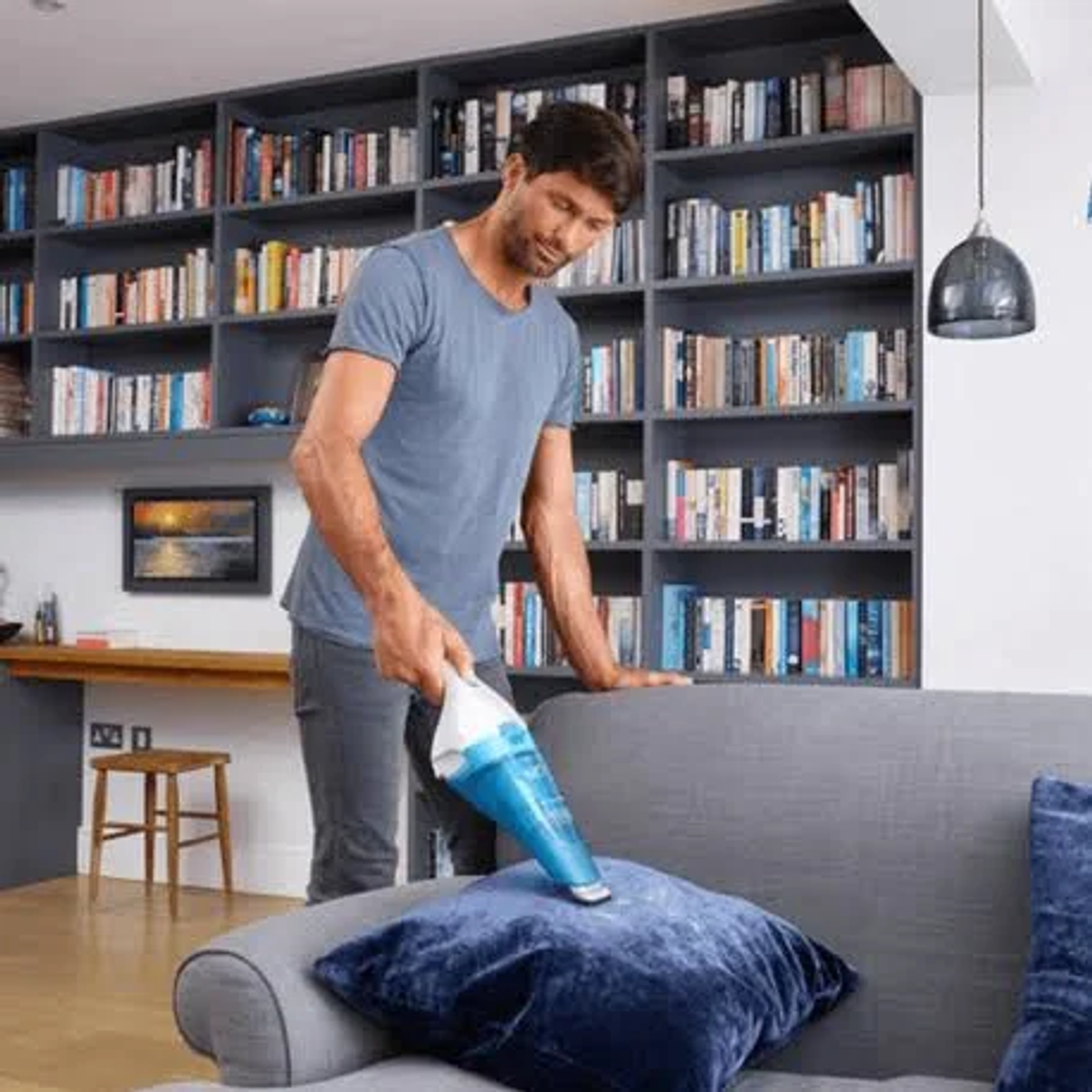 Hombre aspirando una almohada azul en un sofá gris. Estanterías en el fondo.