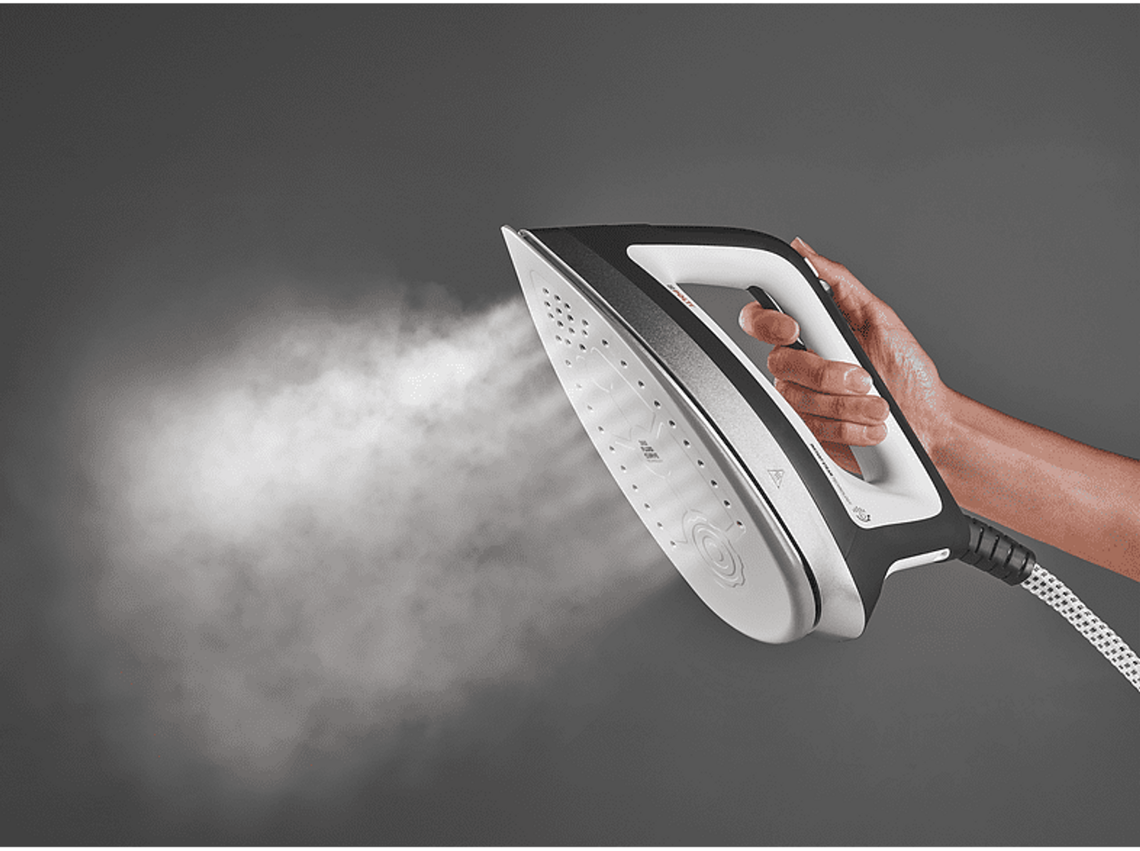 Una mano sosteniendo una plancha de vapor, sale vapor, sobre un fondo gris.