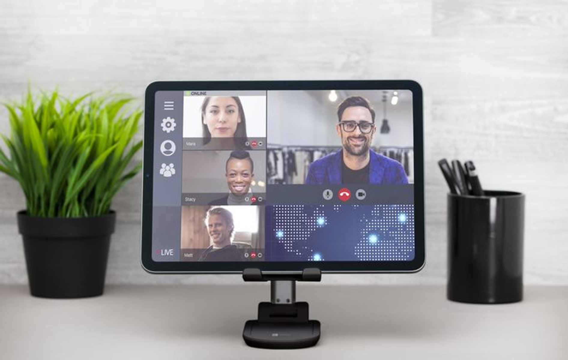 Tableta en un soporte que muestra una videollamada. Personas en la llamada, maceta y portalápices.