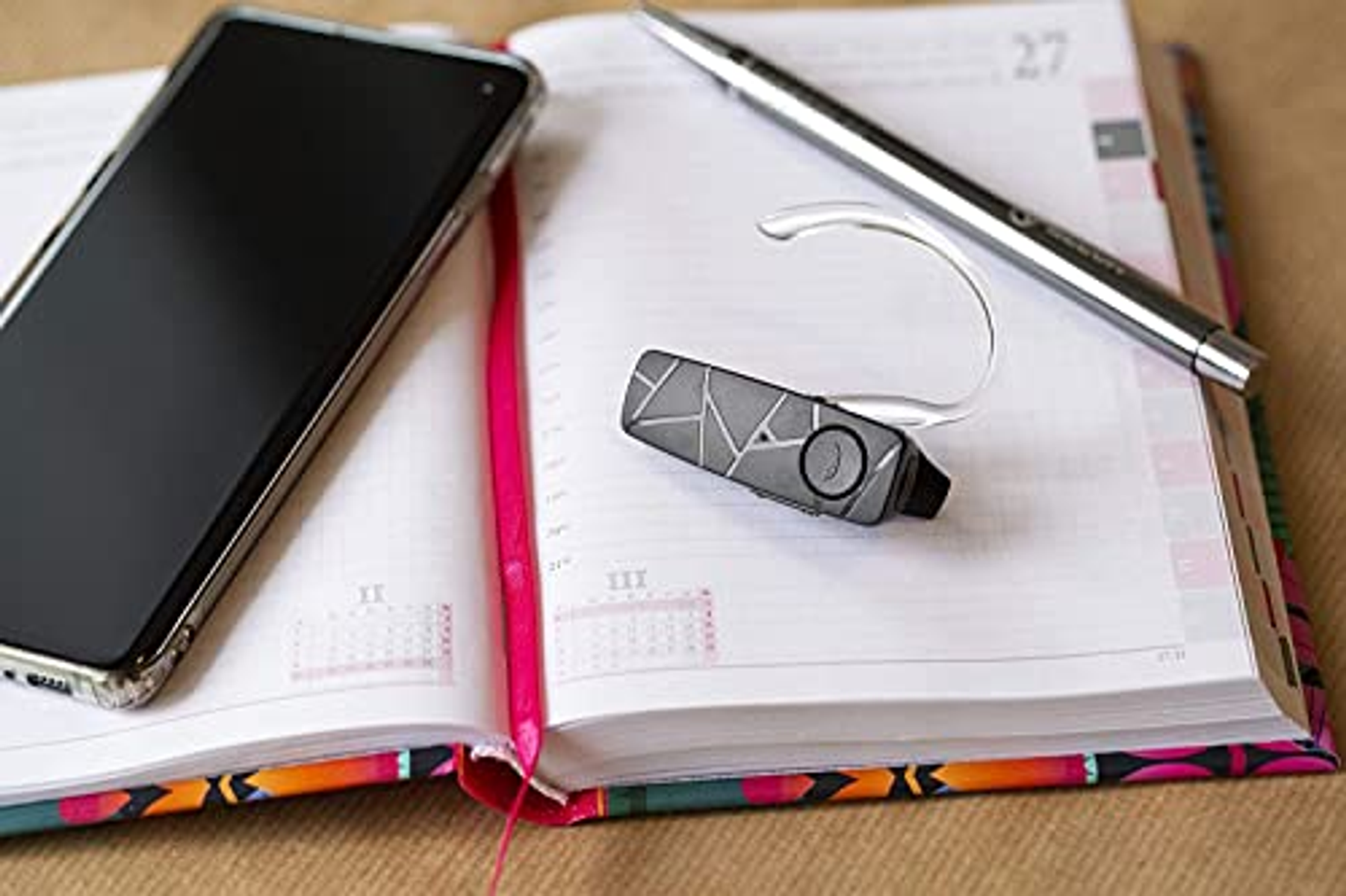 Smartphone, offenes Tagebuch und ein Stift auf dem Schreibtisch. Ein Bluetooth-Headset liegt auf dem Tagebuch.