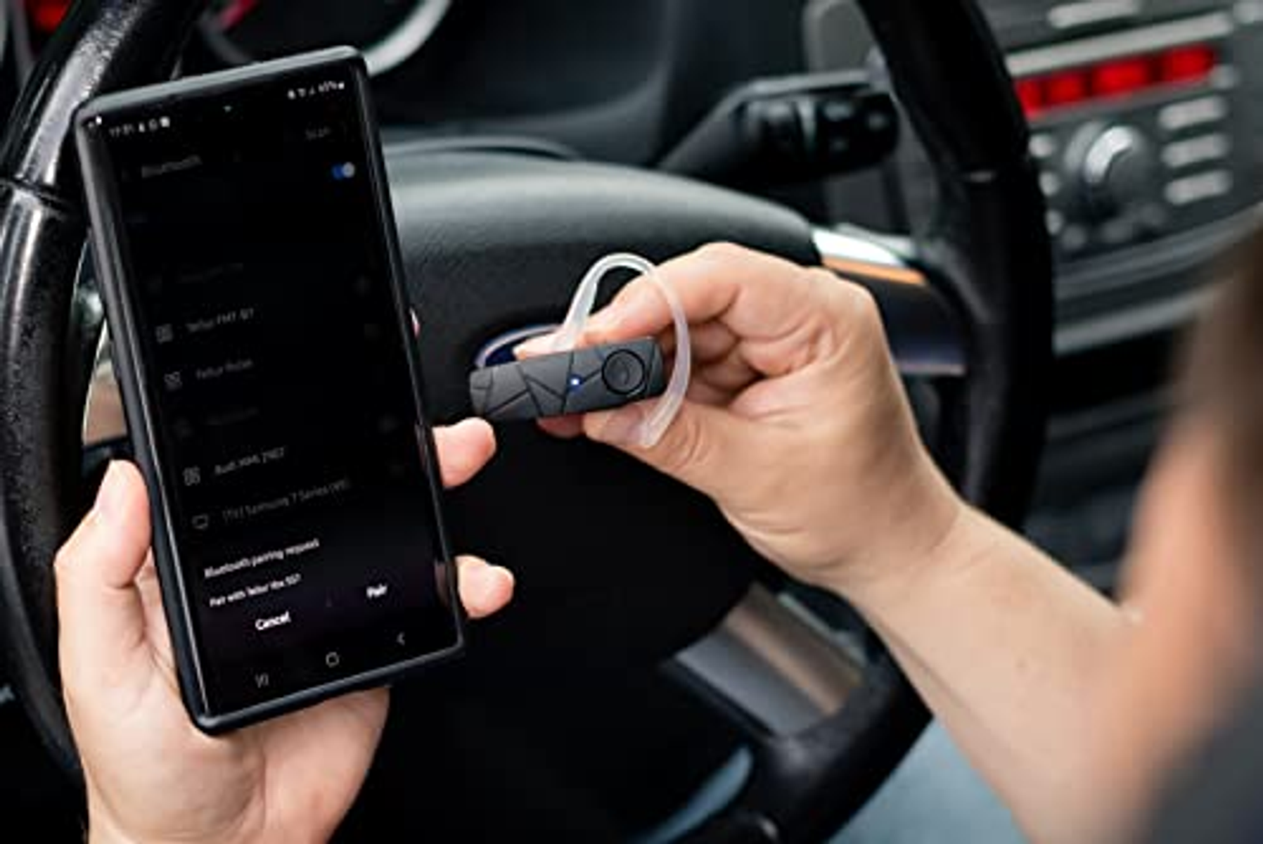 Eine Person, die ein Telefon und ein Bluetooth-Headset in einem Auto hält, neben einem Lenkrad.