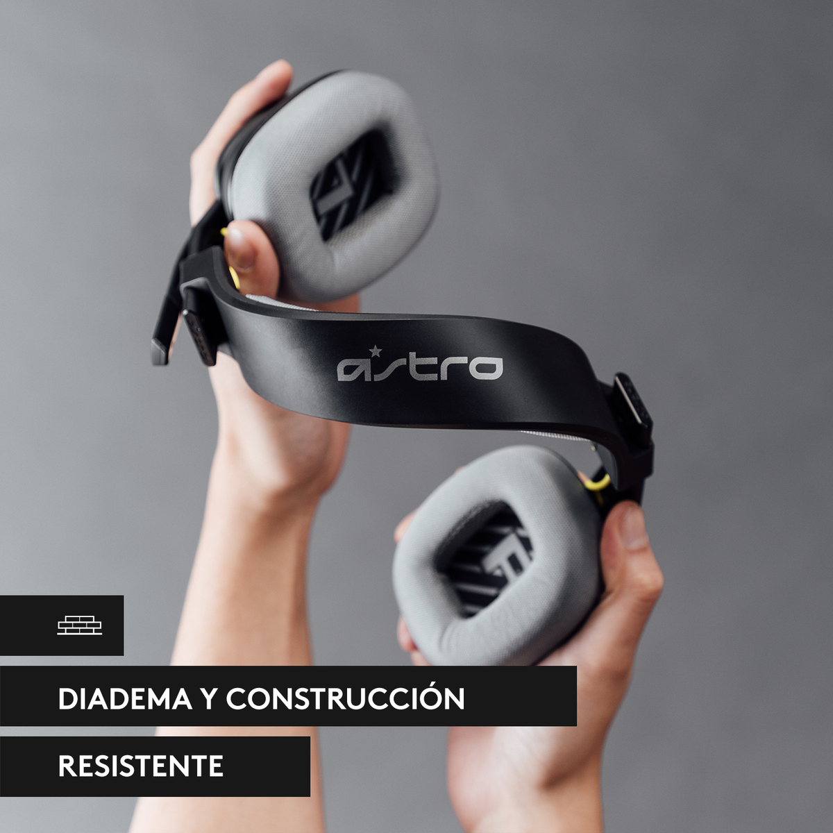 Ręce trzymające czarno-szary zestaw słuchawkowy Astro. Tekst brzmi „DIADEMA Y CONSTRUCCIÓN RESISTENTE”.