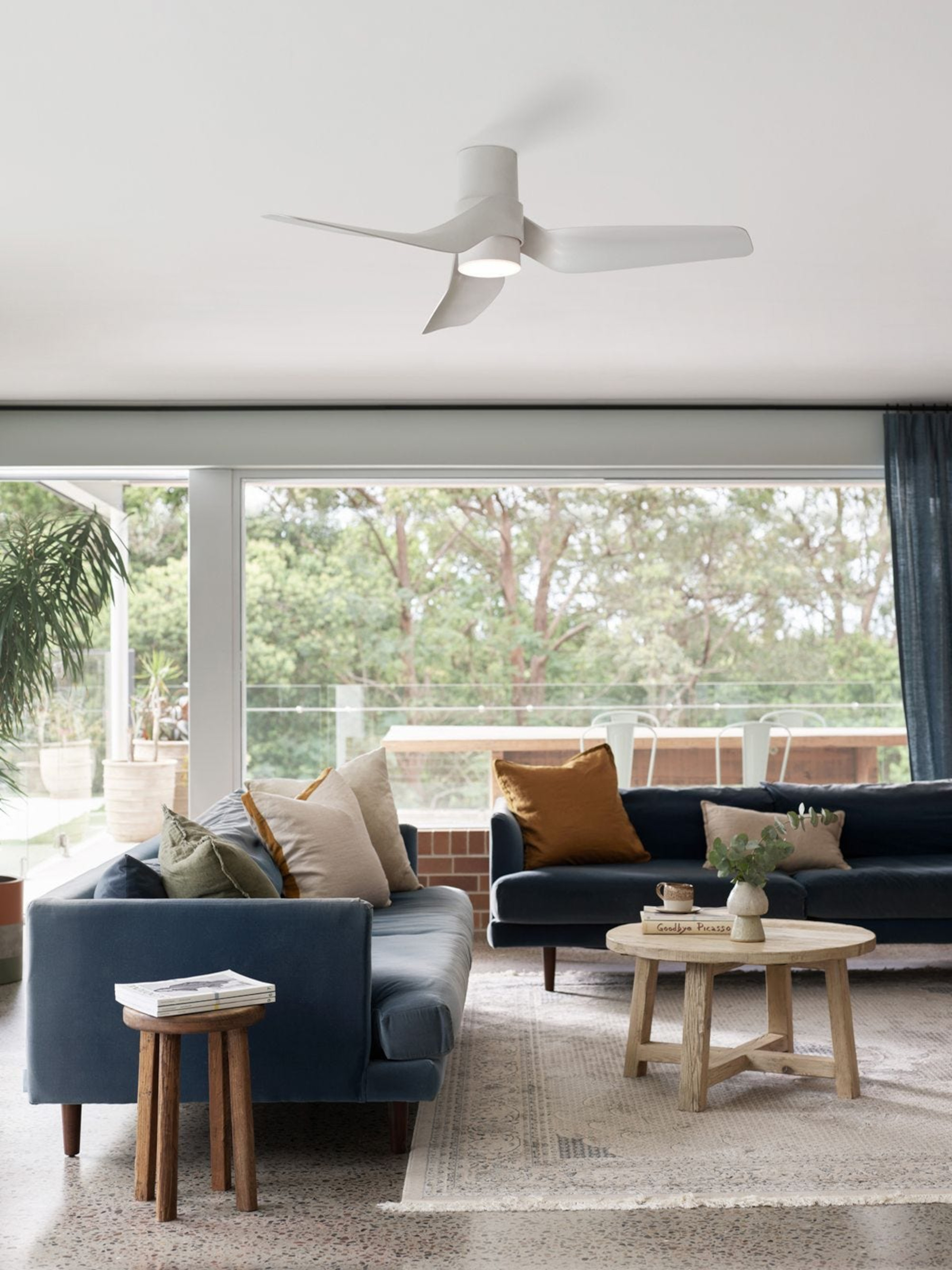 Wohnzimmer mit blauen Sofas, Deckenventilator und großen Fenstern mit Blick auf Bäume.