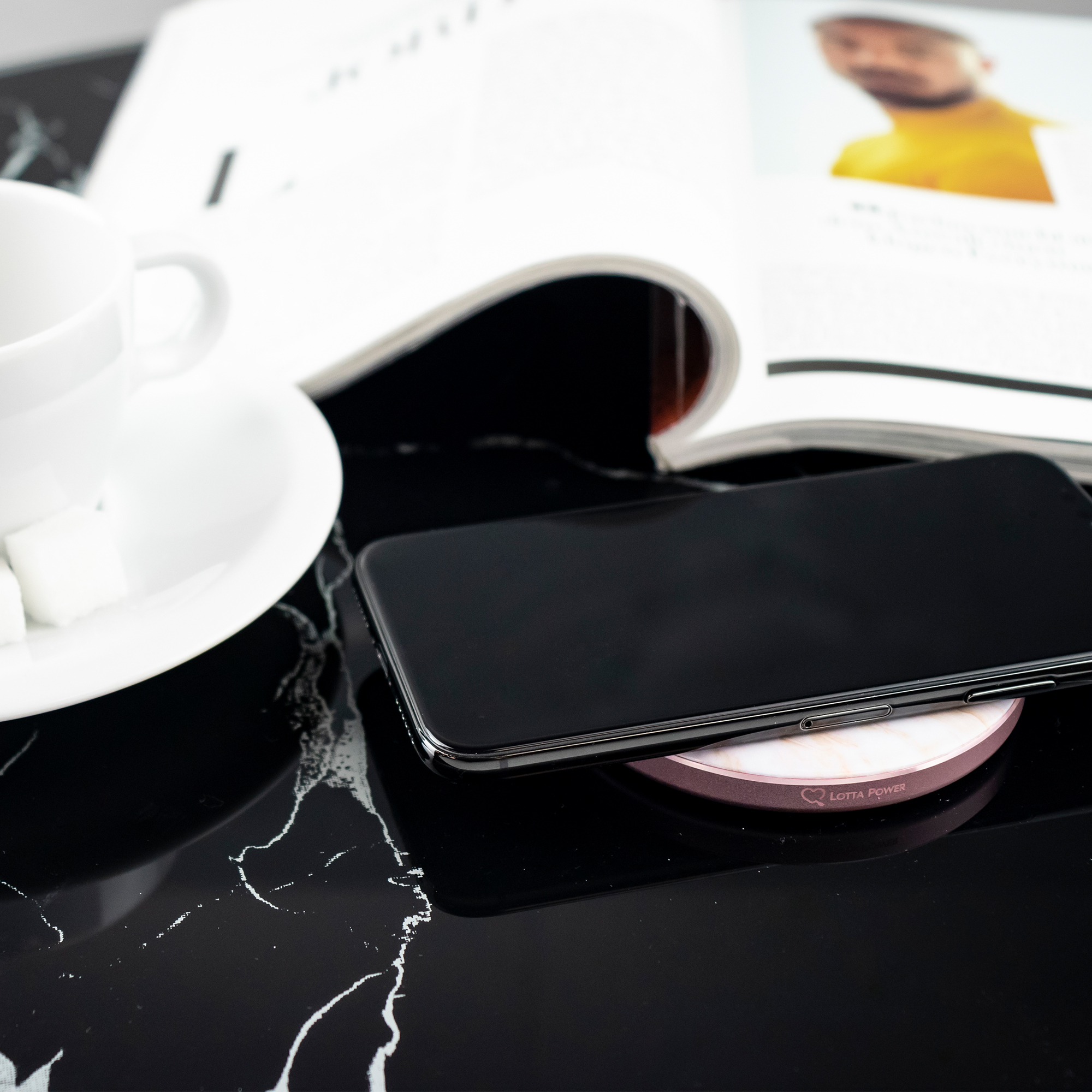 Schwarzes Telefon auf kabellosem Ladegerät, mit Zeitschrift und Kaffee.