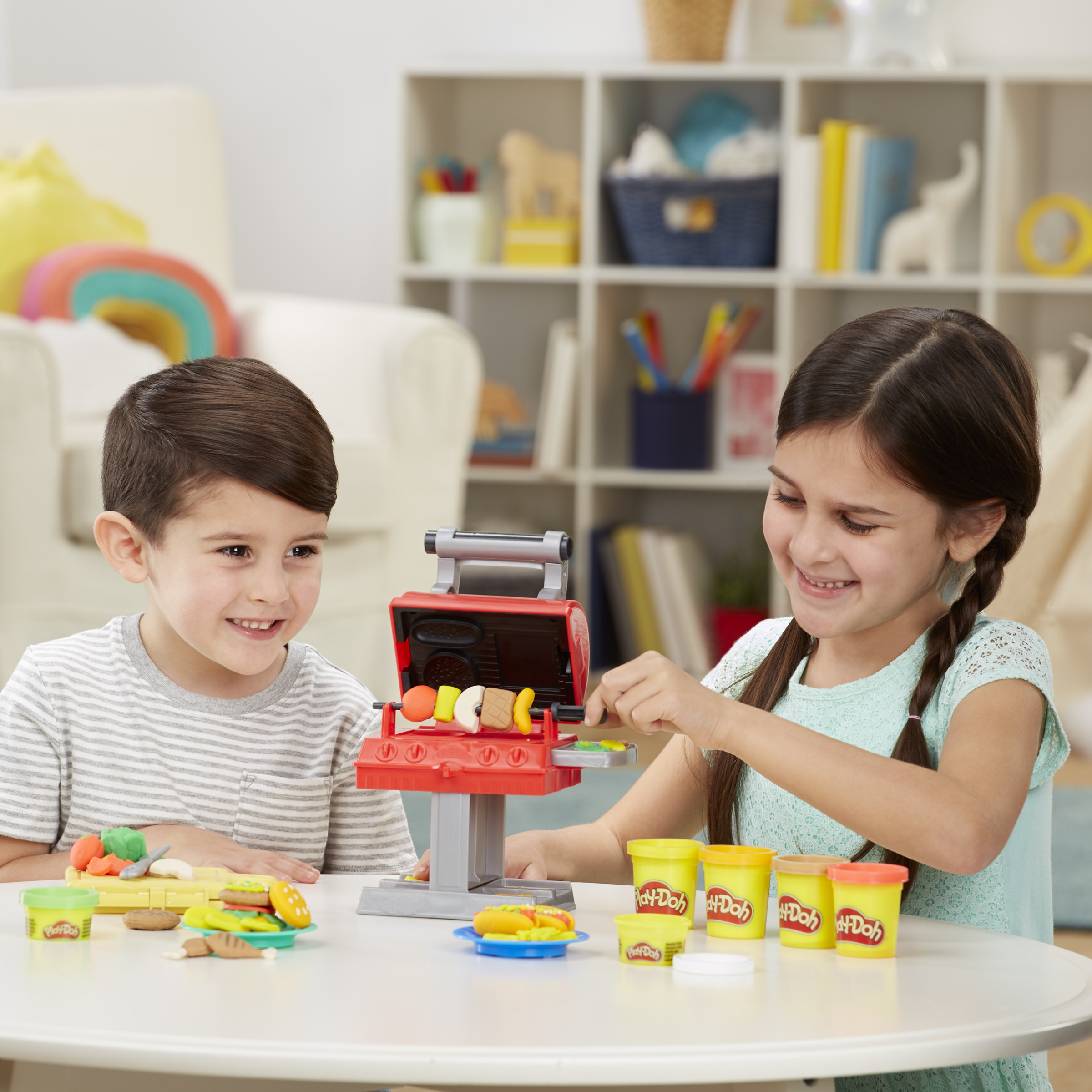 Dos niños jugando con una parrilla de juguete. Una niña pone comida de juguete. Latas de Play-Doh en la mesa.