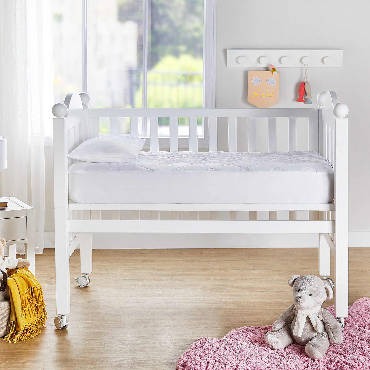 Una cuna blanca con colchón y almohada. Hay una alfombra rosa y un oso de peluche en el suelo.