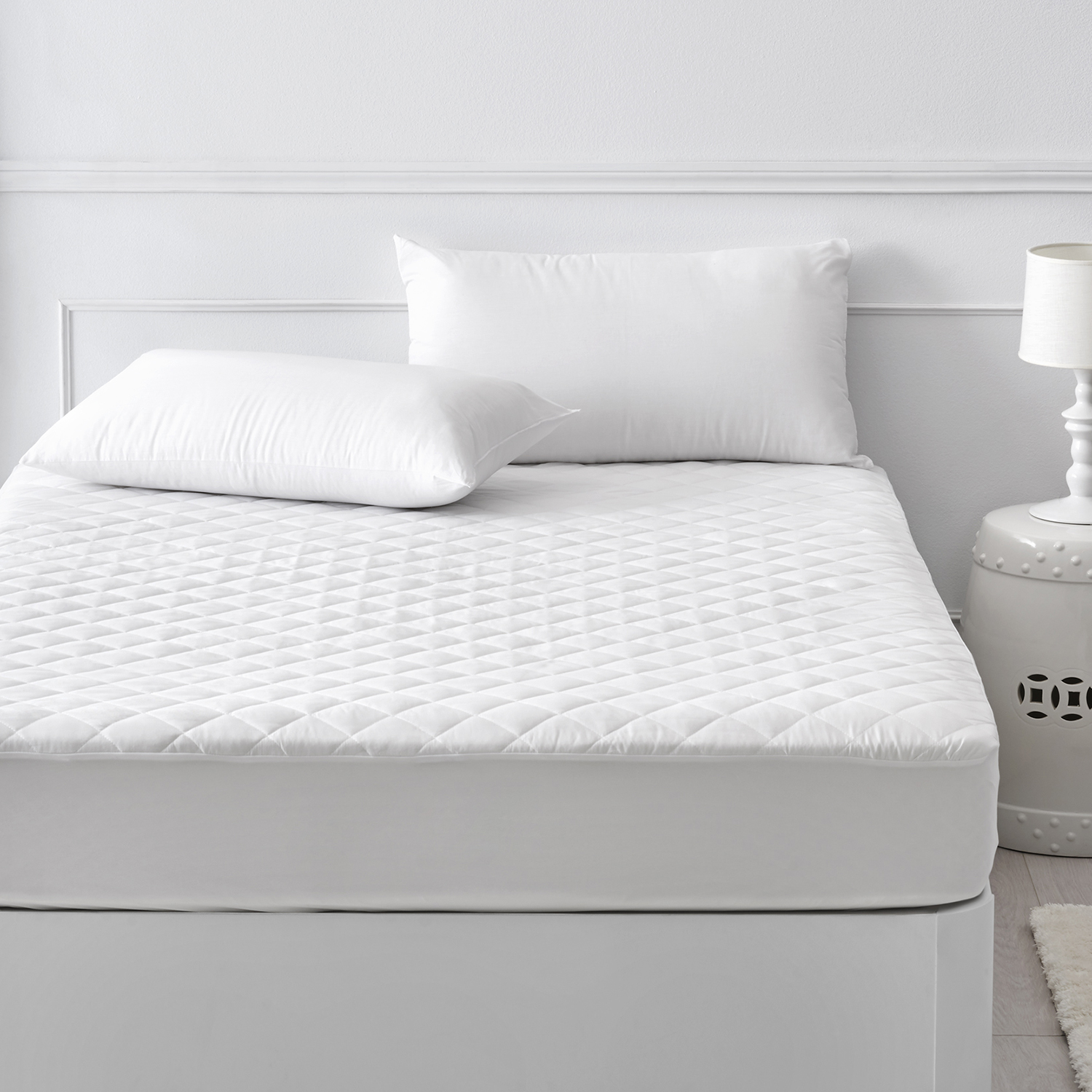 Una cama con un cubrecolchón acolchado blanco, dos almohadas y una lámpara blanca sobre un taburete.