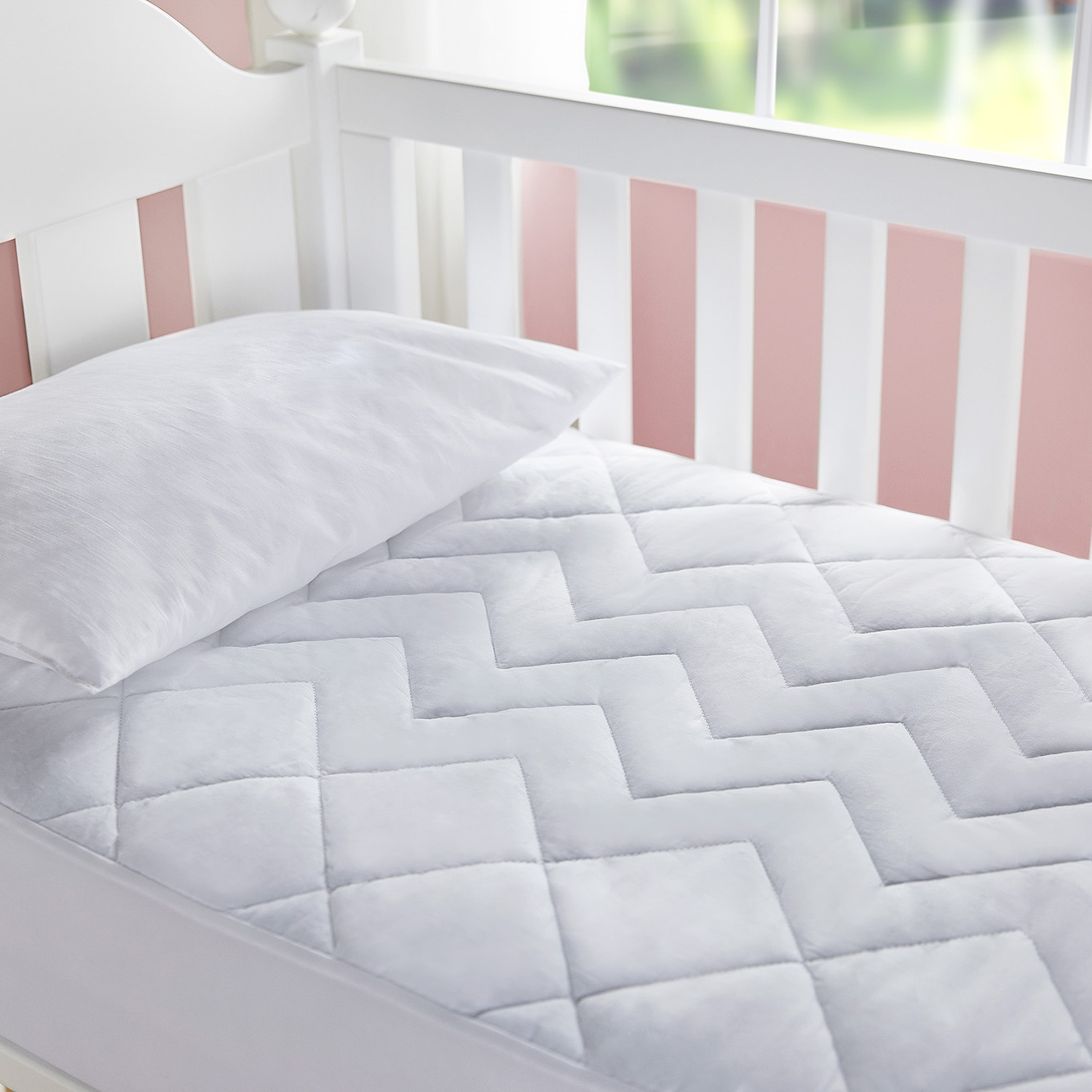 Una cuna con una almohada blanca y un colchón acolchado blanco. La pared es de rayas rosas y blancas.
