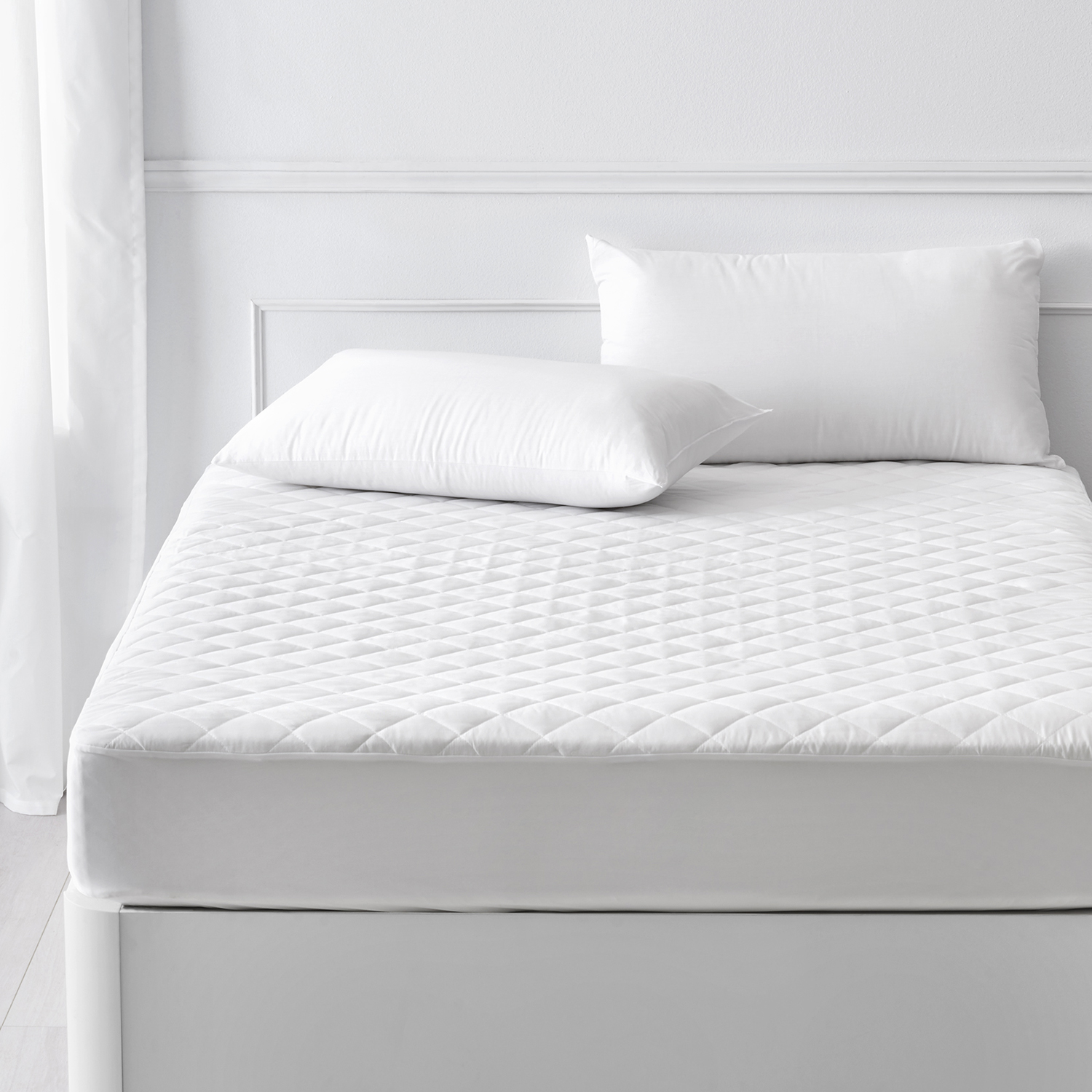 Cama blanca con una funda de colchón acolchada y dos almohadas blancas. La cama está contra una pared blanca.