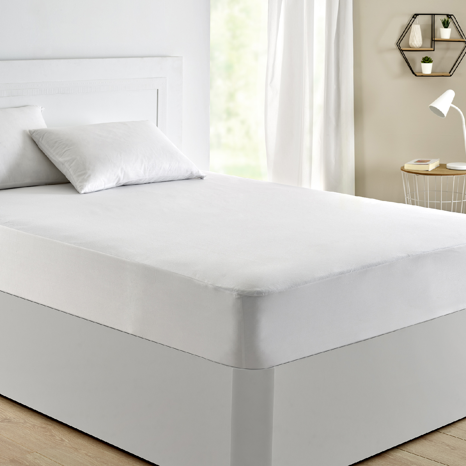 Cama blanca con ropa de cama blanca. Se ven una mesita de noche y una lámpara. Una ventana con cortinas al fondo.