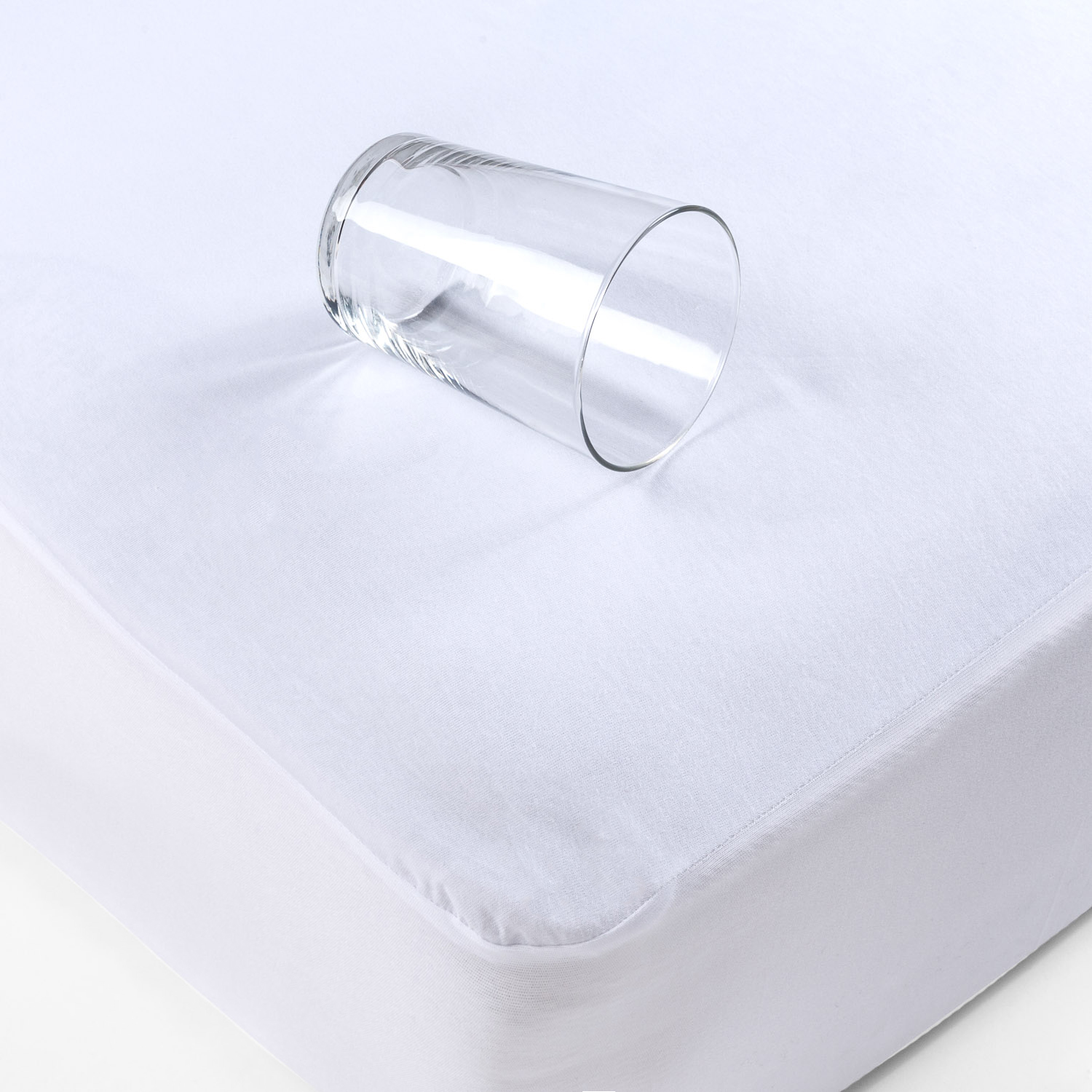 Un vaso transparente sobre un protector de colchón blanco, lateral.