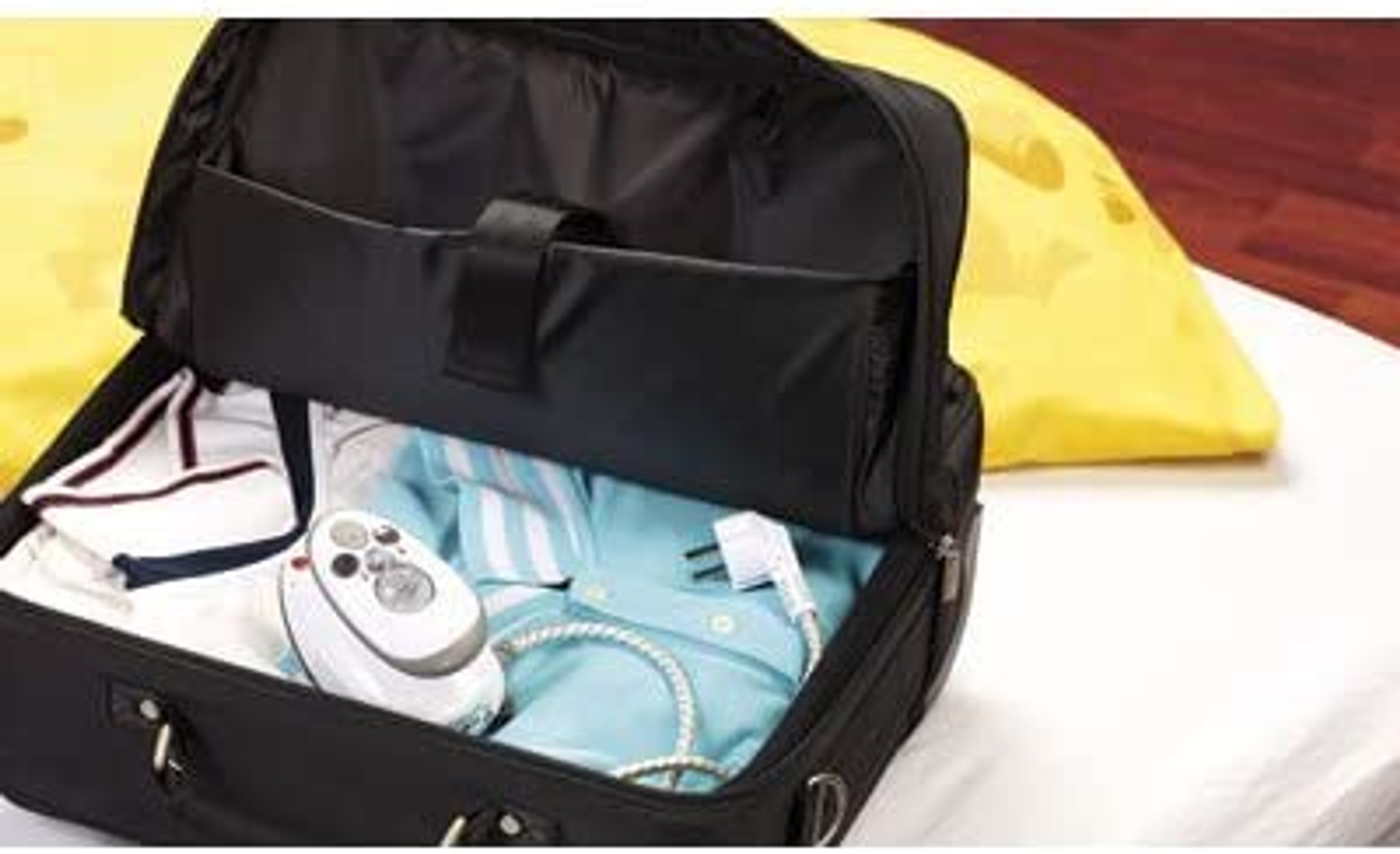 Schwarze Reisetasche mit Kleidung und Bügeleisen. Gelber Kissenhintergrund.