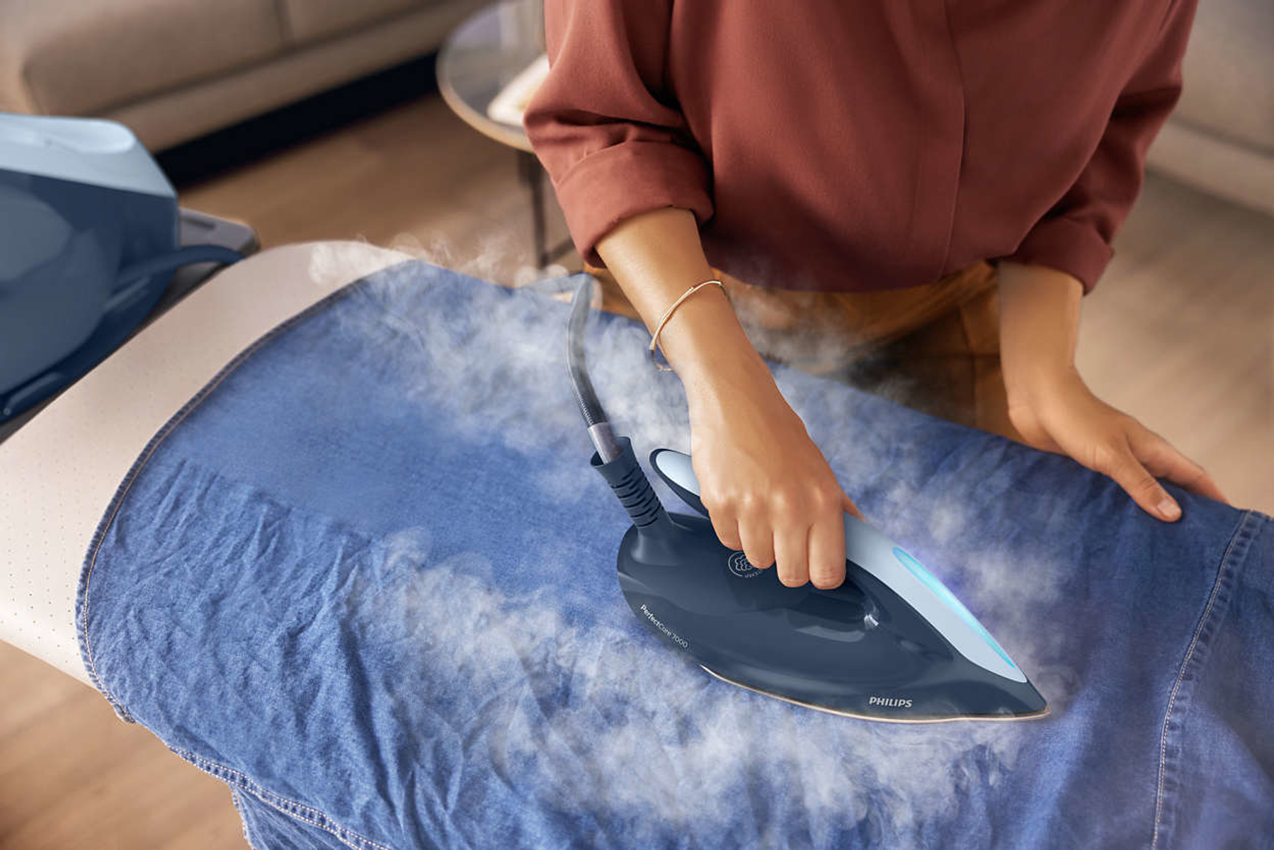 Donna che stira una camicia di jeans blu su un'asse da stiro con un ferro. Vapore visibile.