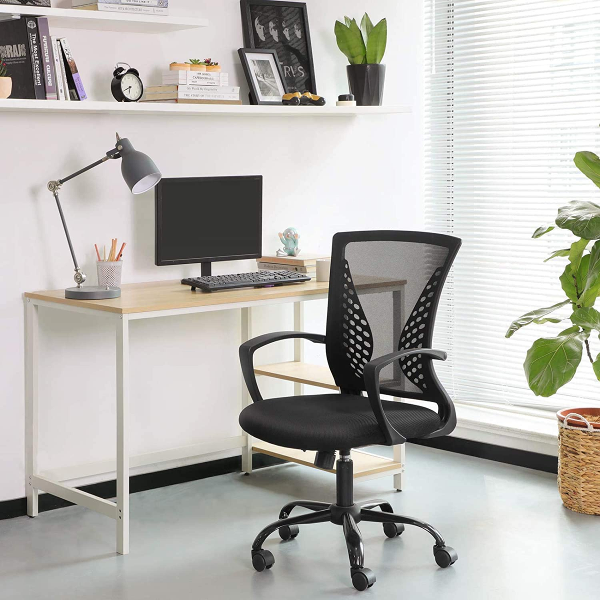 Silla de oficina negra con malla en un escritorio. Monitor, lámpara y planta visibles.
