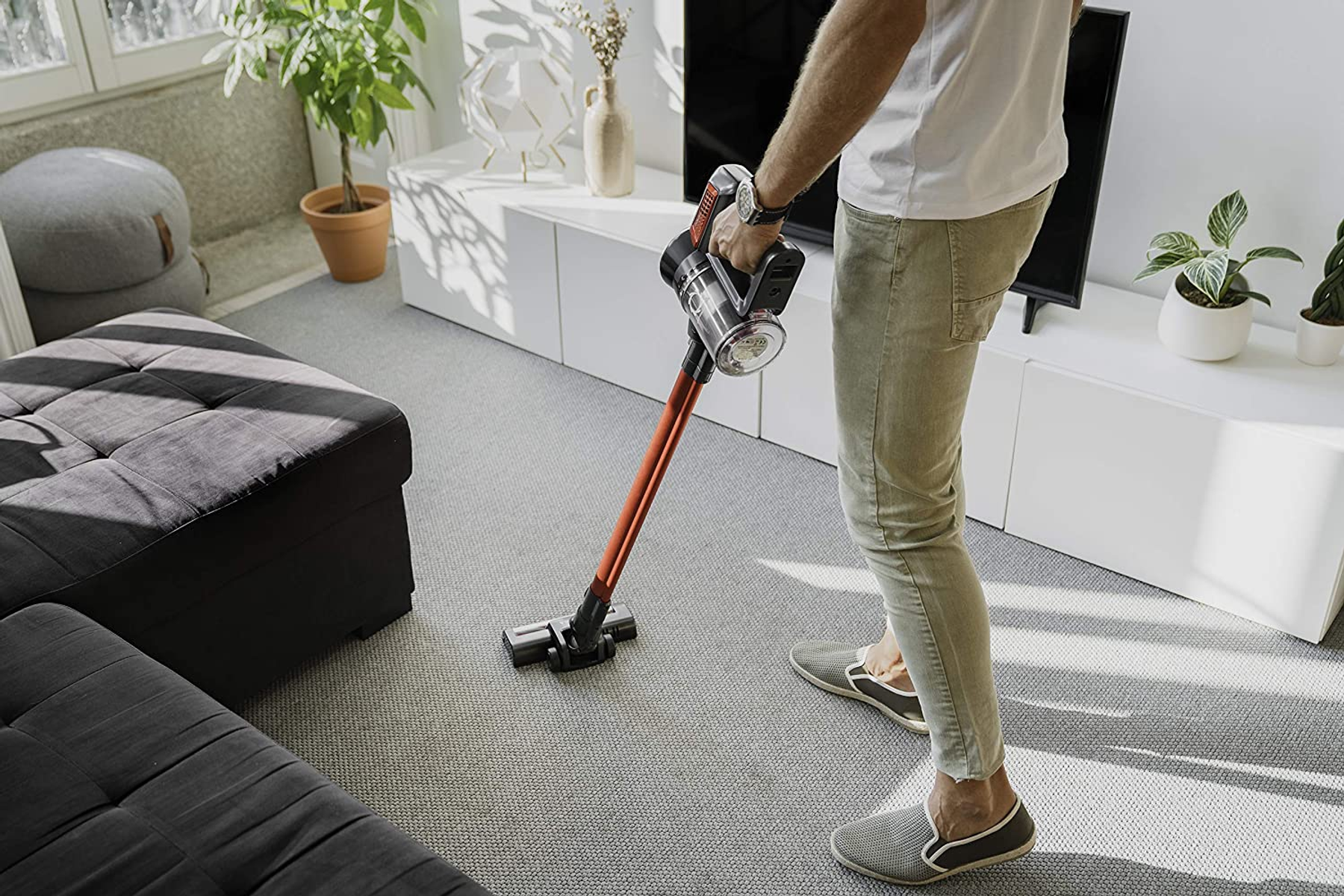 Una persona aspirando un piso alfombrado gris con una aspiradora roja y negra.
