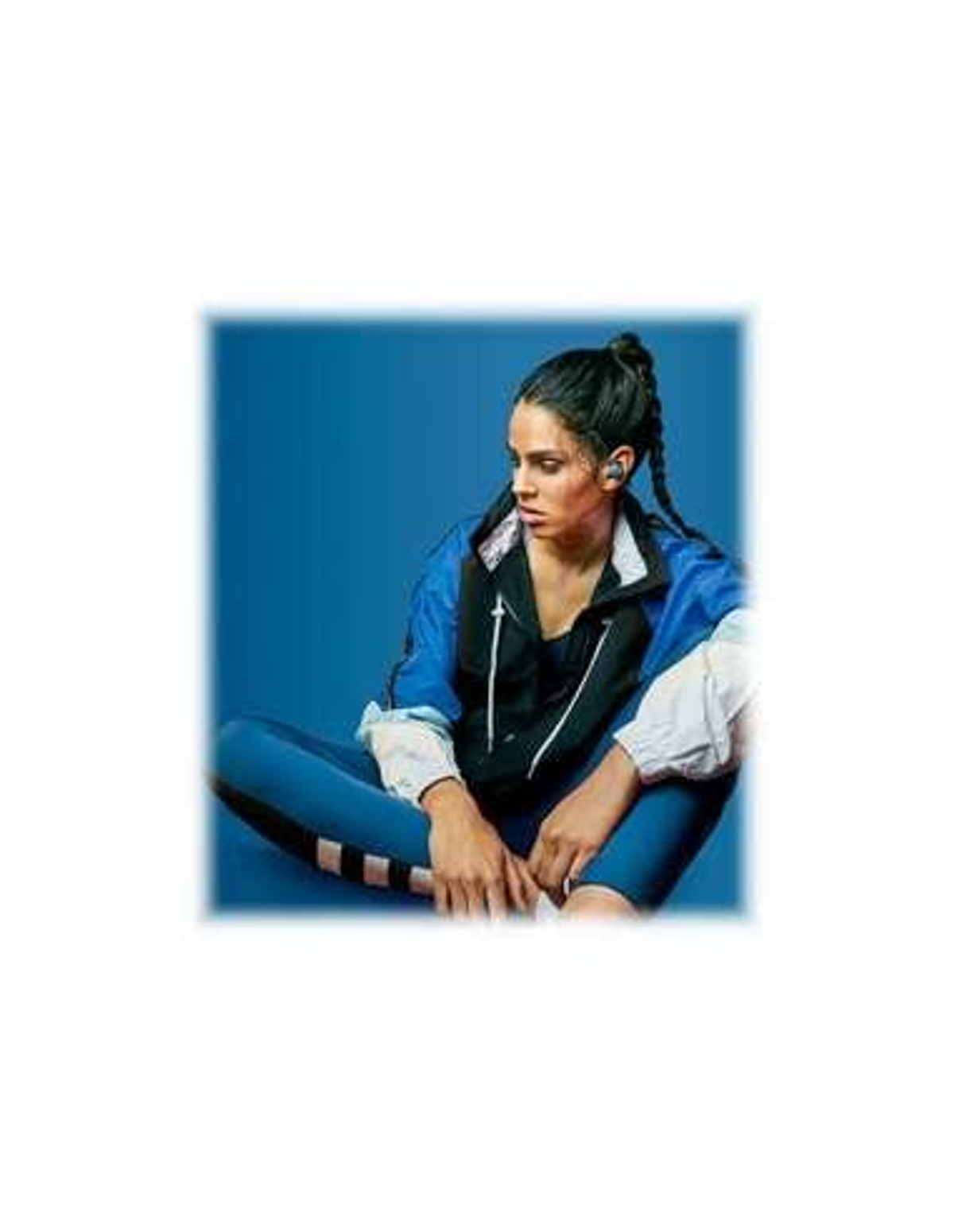 Frau in blauer Jacke und Leggings, trägt Ohrhörer, sitzt mit gekreuzten Beinen vor blauem Hintergrund.