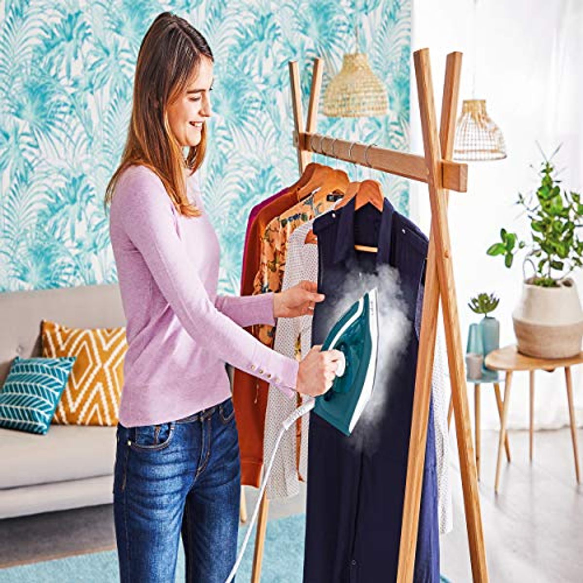 Mujer planchando camisa azul marino en perchero de madera, vapor visible. Papel pintado floral, sofá y planta detrás.