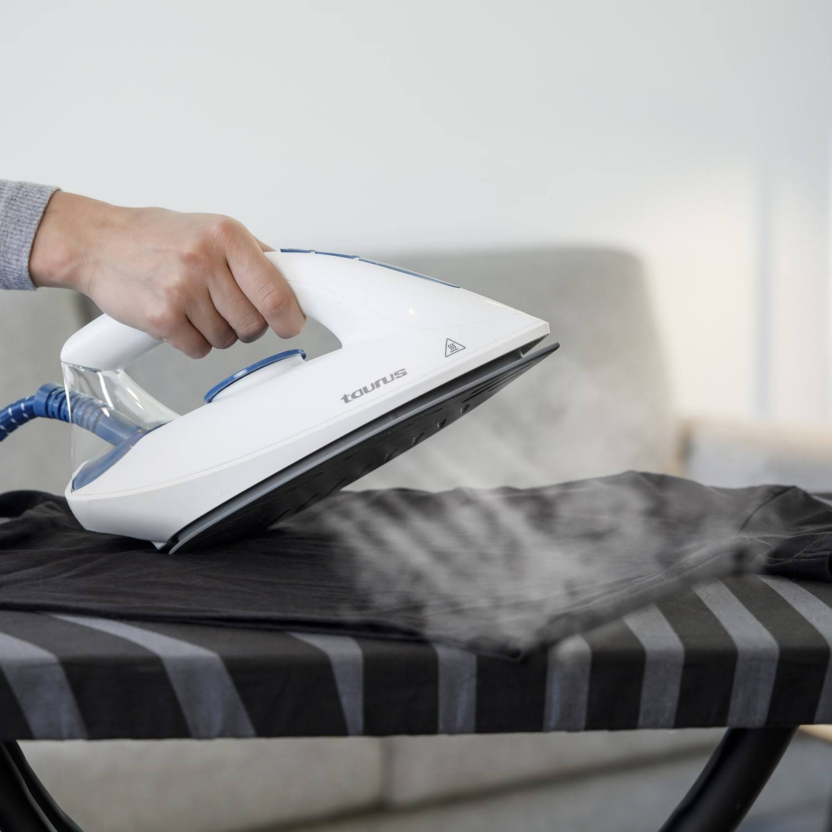 Una plancha blanca y azul que vaporiza una camisa negra sobre una tabla de planchar a rayas.