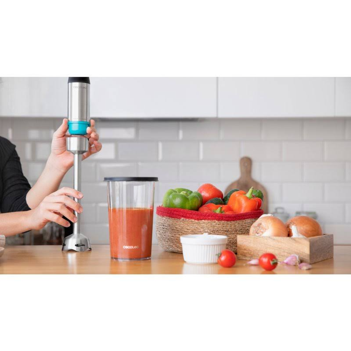 Een hand die een zilveren staafmixer vasthoudt naast een glas tomatensoep en ingrediënten.