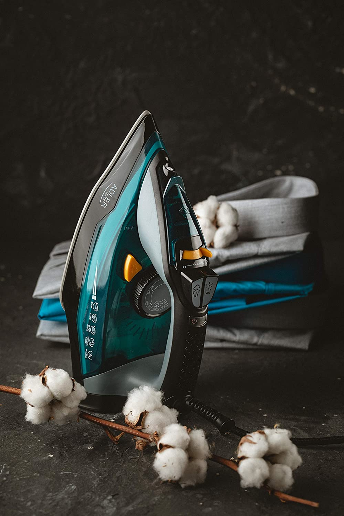 Żelazko na ciemnej powierzchni obok kwiatów bawełny i złożonych ubrań. Żelazko jest turkusowe i czarne.