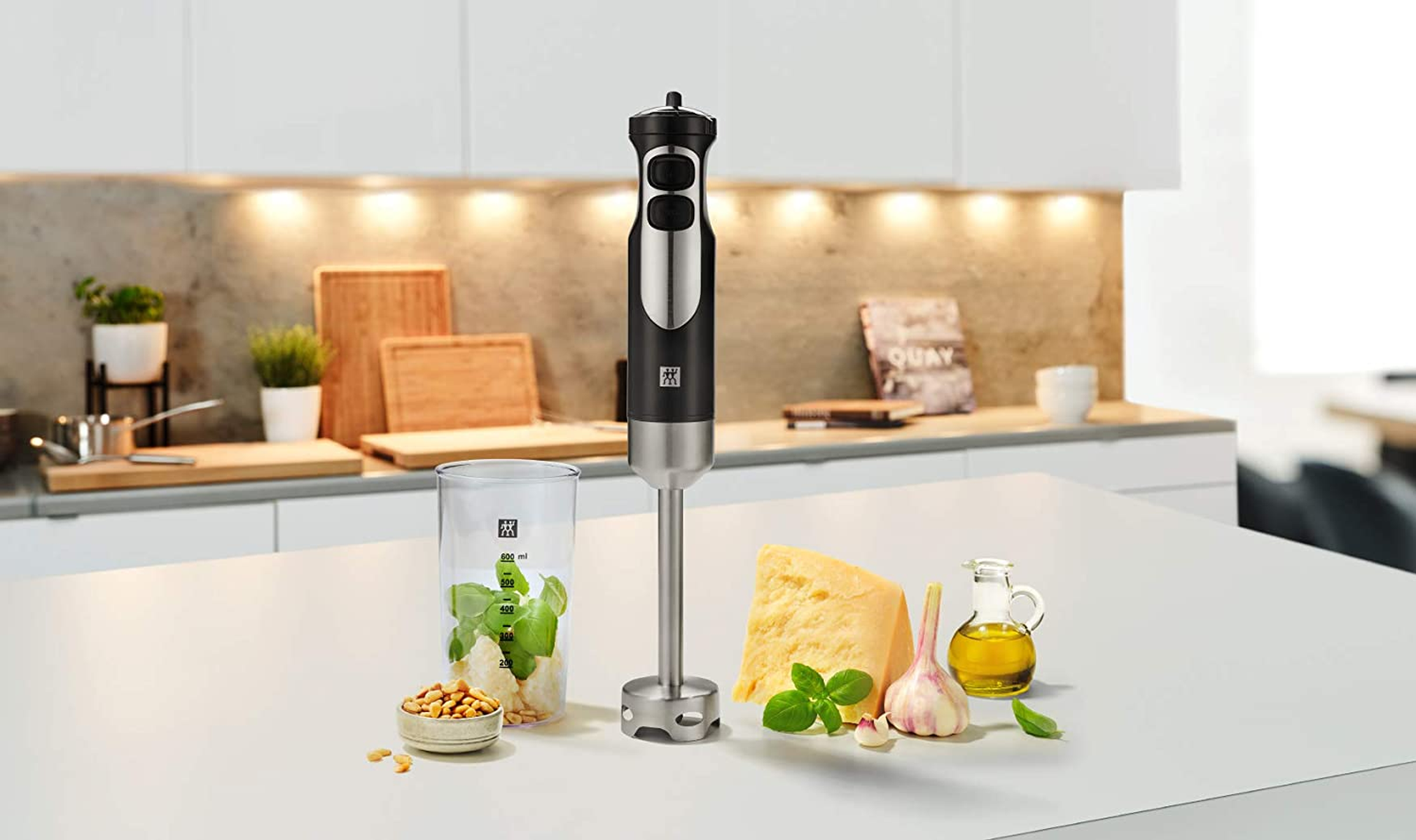 Frullatore a immersione con accessori e ingredienti su un piano bianco.