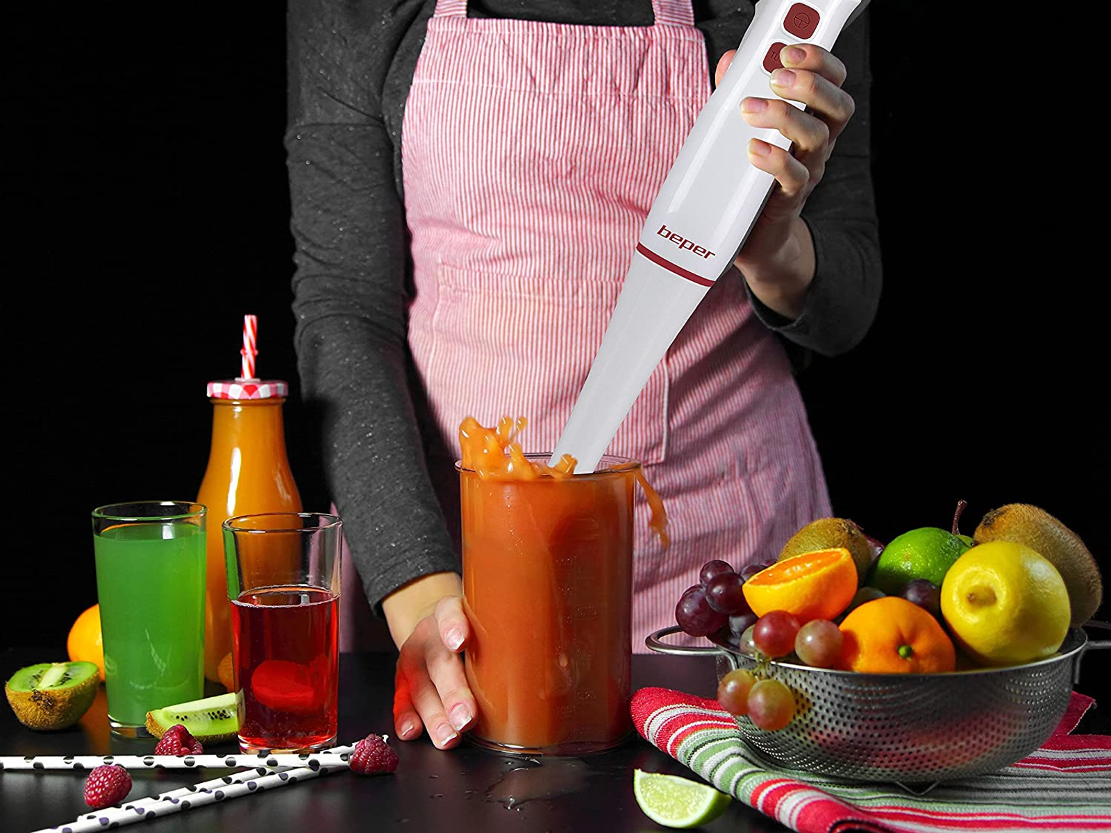 Una persona usa un frullatore a immersione per frullare una bevanda in un bicchiere. Frutta e succhi sono sul tavolo.