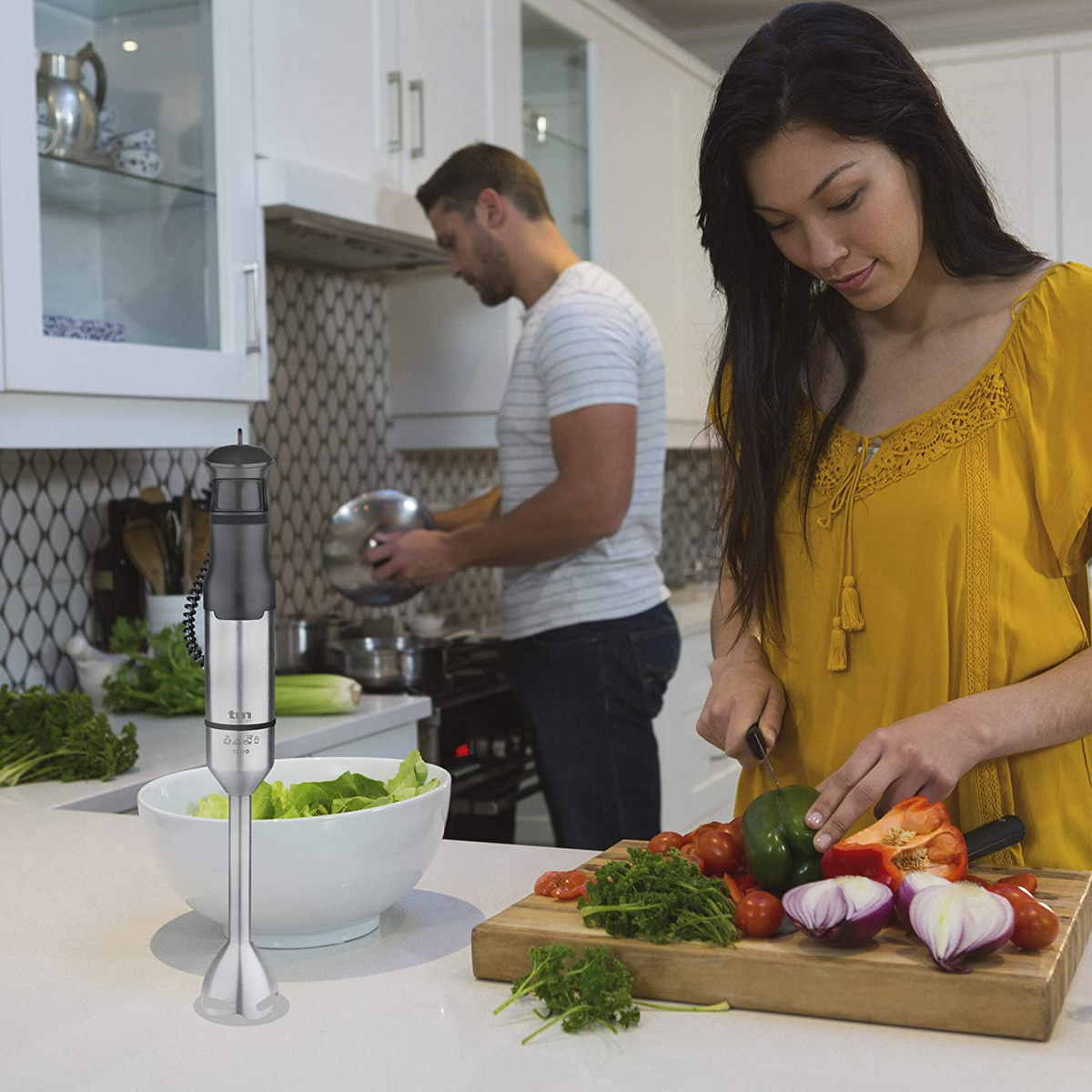 Una mujer con camisa amarilla corta verduras en una cocina con un hombre en el fondo.