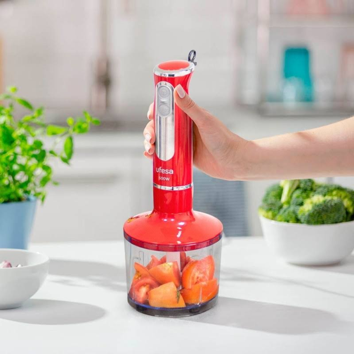 Mano di donna che tiene un frullatore a immersione rosso pieno di pomodori tritati. Broccoli sullo sfondo.