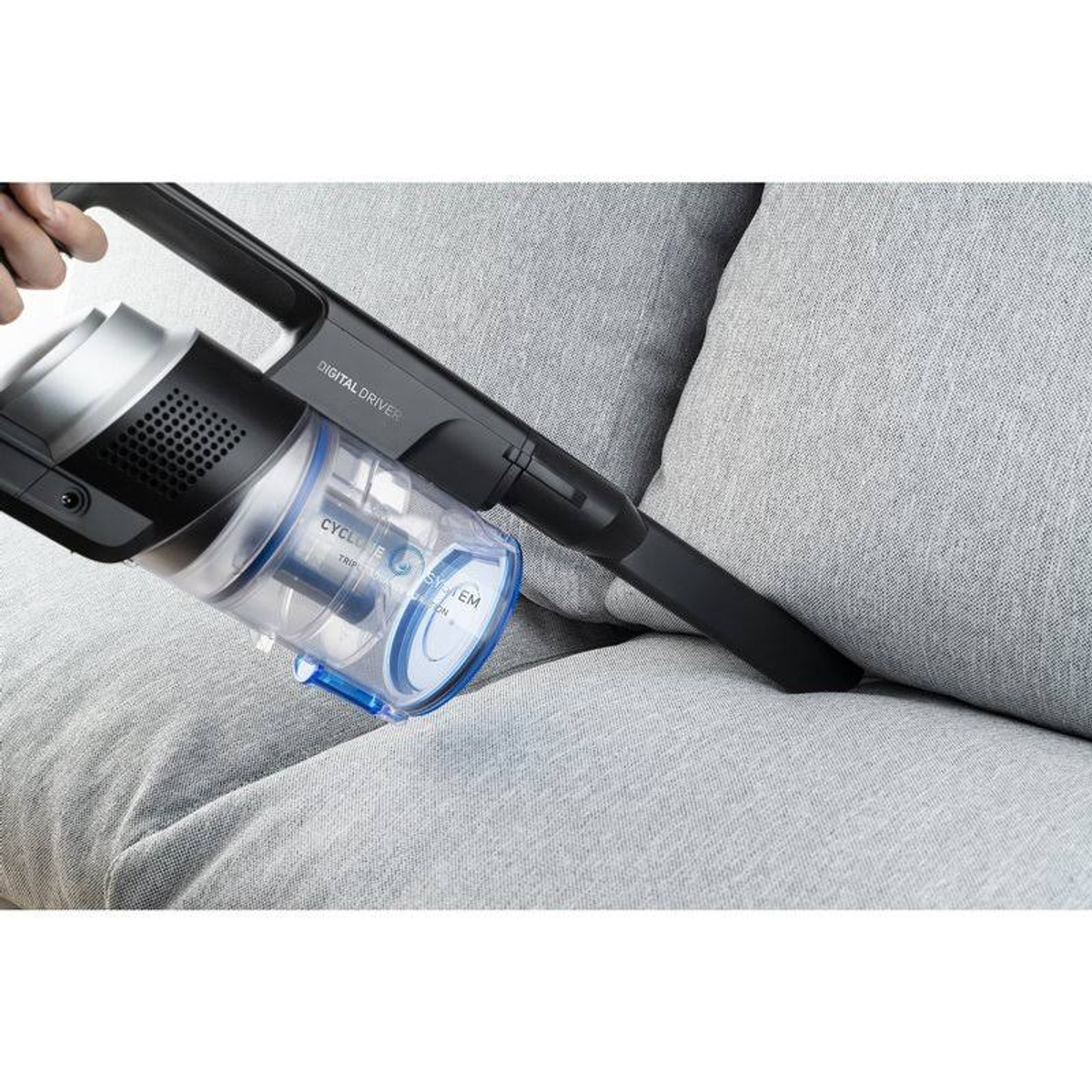 Eine Person saugt ein graues Sofa mit einem kabellosen Staubsauger. Der Staubsauger hat einen blauen Behälter.