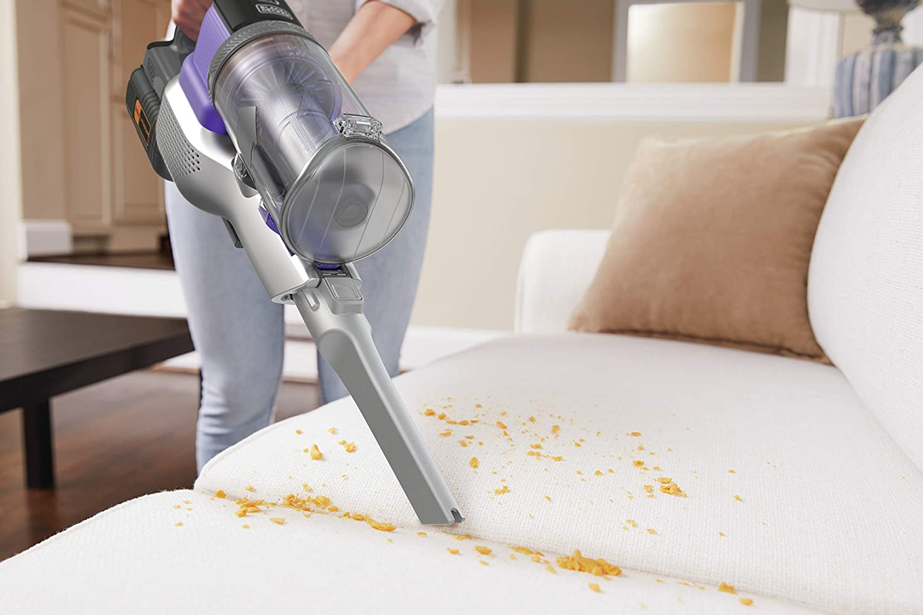 A person vacuuming a white couch with a cordless vacuum. Food crumbs are visible.