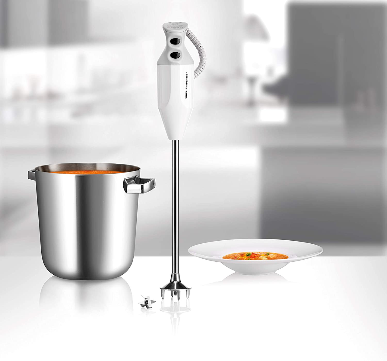 Ein weißer Stabmixer in Gebrauch, der Suppe in einem großen Metalltopf püriert. Eine Schüssel Suppe steht daneben.