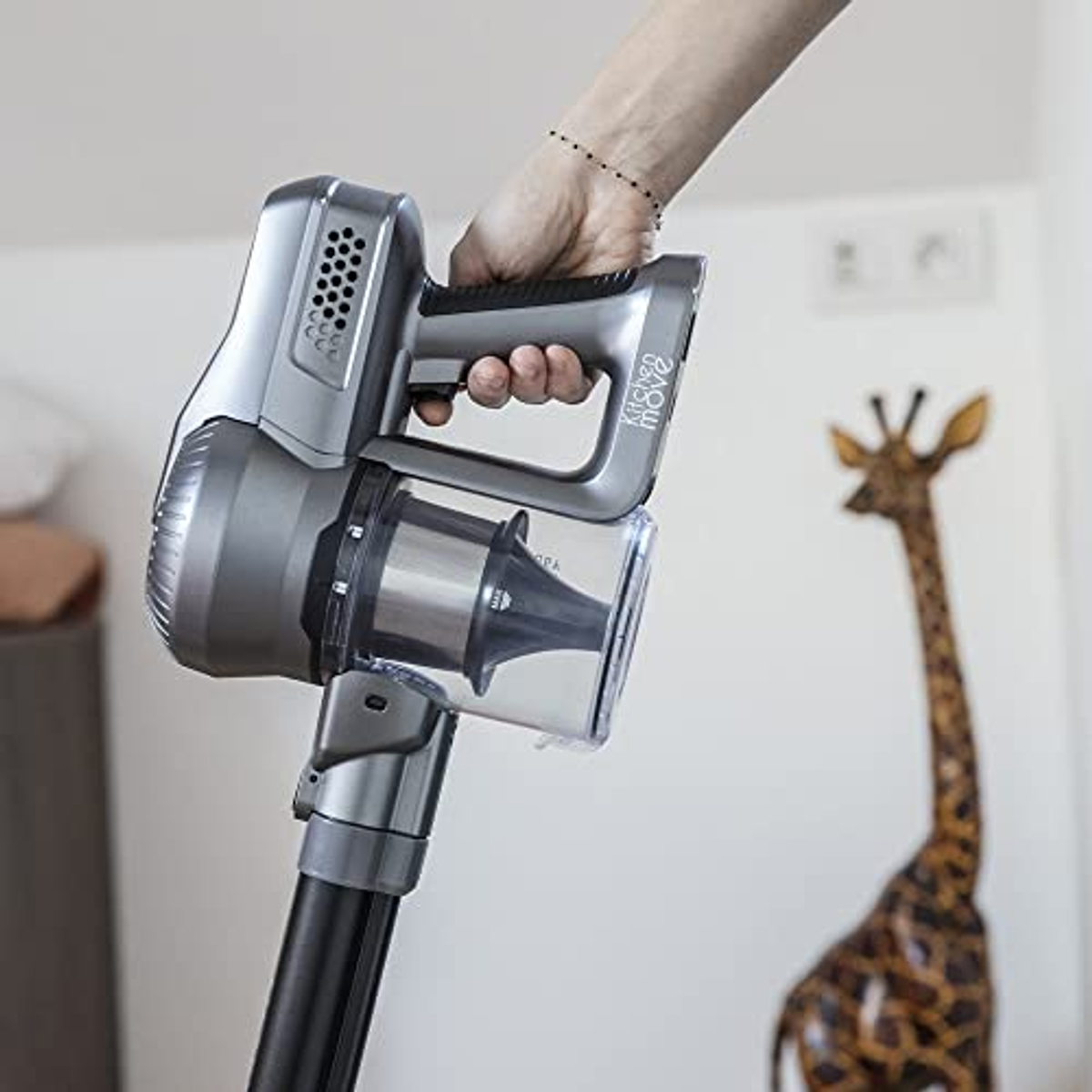 Hand hält silbernen Staubsauger, Giraffen-Skulptur im Hintergrund.