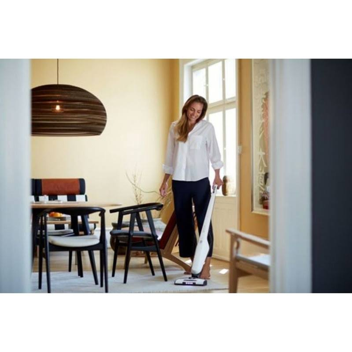 Een vrouw die een kamer met houten meubels stofzuigt. Ze glimlacht en draagt een wit overhemd.