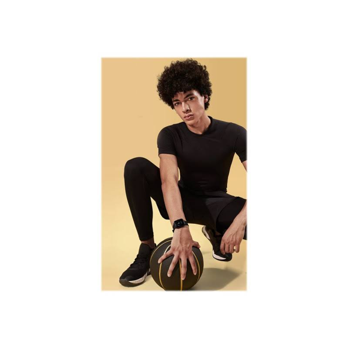 Un uomo in abbigliamento sportivo nero si accovaccia, tenendo un pallone da basket, indossando uno smartwatch.