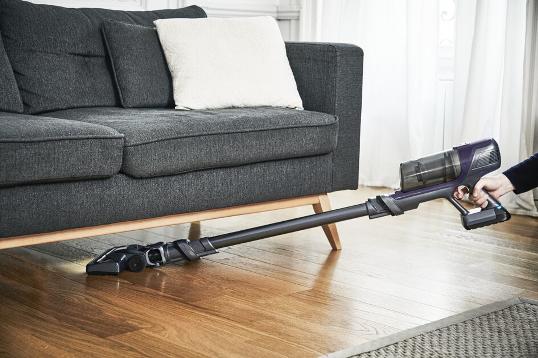 Persona aspirando un piso de madera con una aspiradora inalámbrica. Sofá gris visible en el fondo.