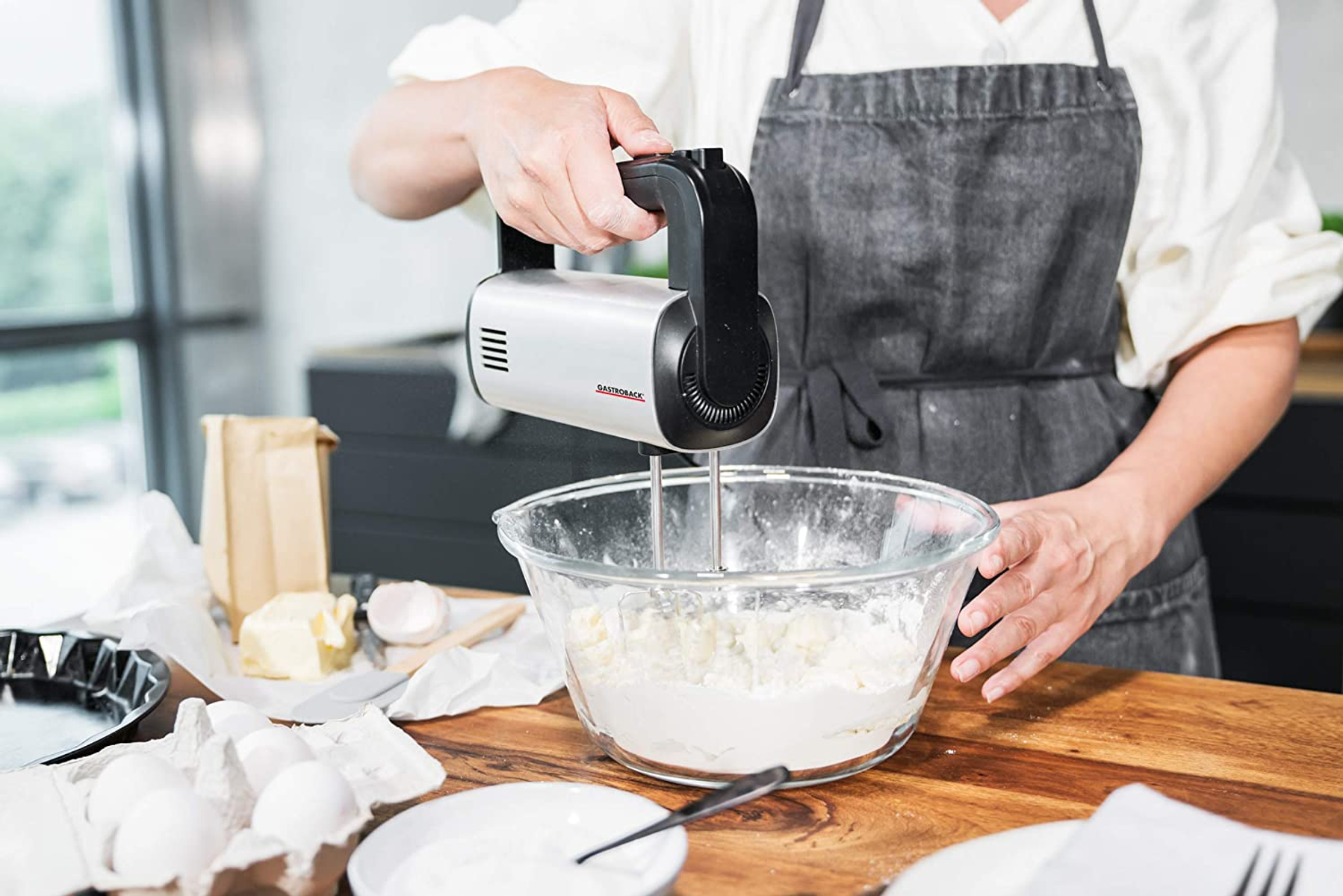 Donna che usa un frullatore elettrico per mescolare gli ingredienti in una ciotola di vetro su un bancone di legno.