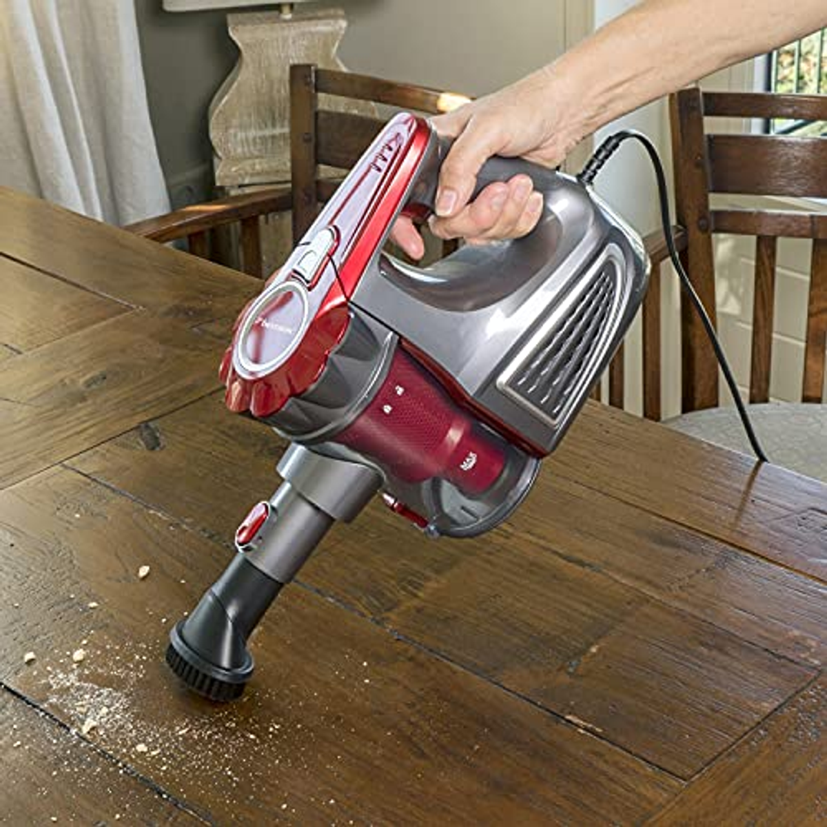 Hand hält einen rot-grauen Staubsauger, der einen Holztisch mit Krümeln reinigt.