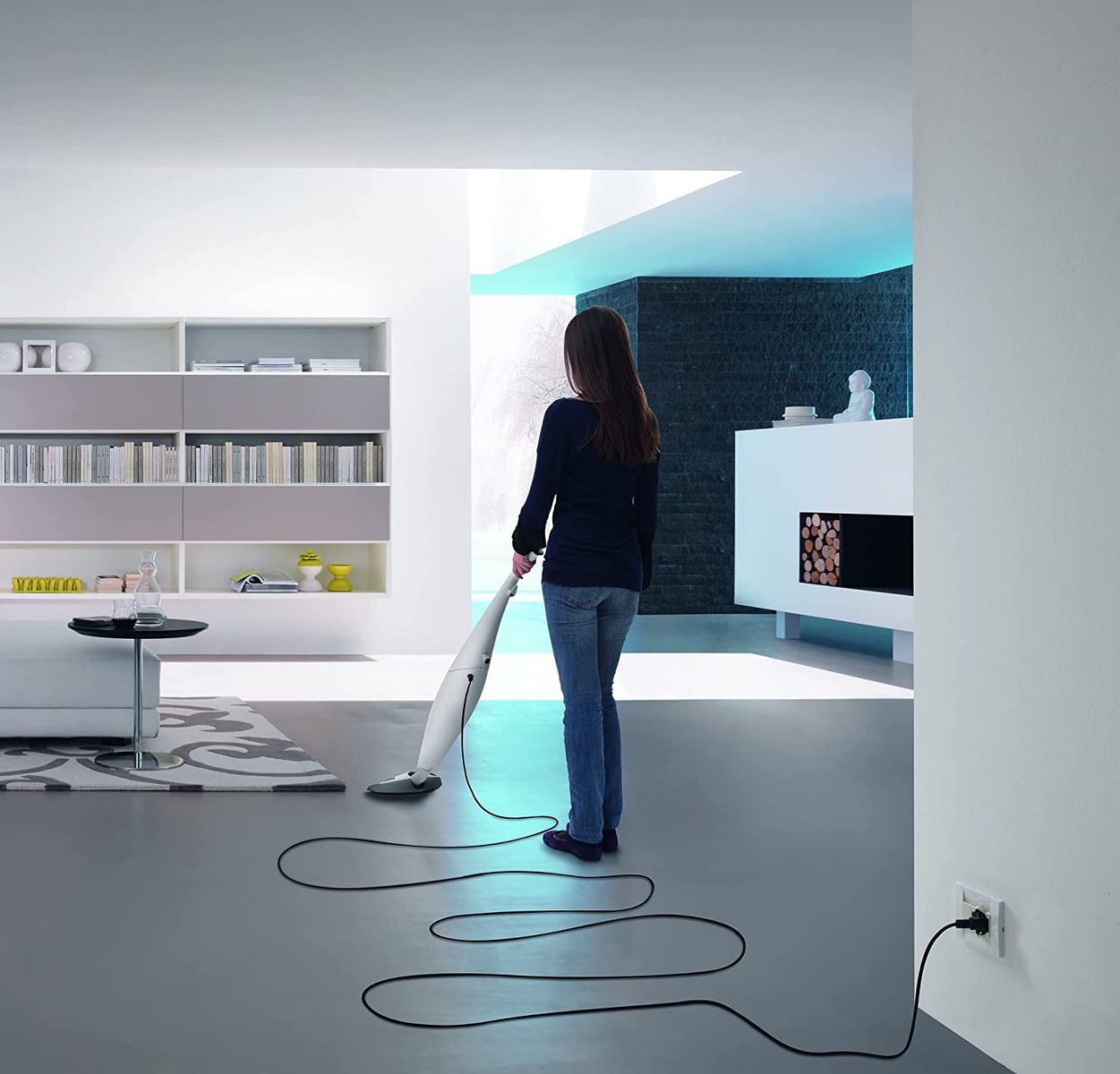 Kobieta odkurzająca nowoczesny salon z długim, pętlującym kablem i wtyczką w ścianie.