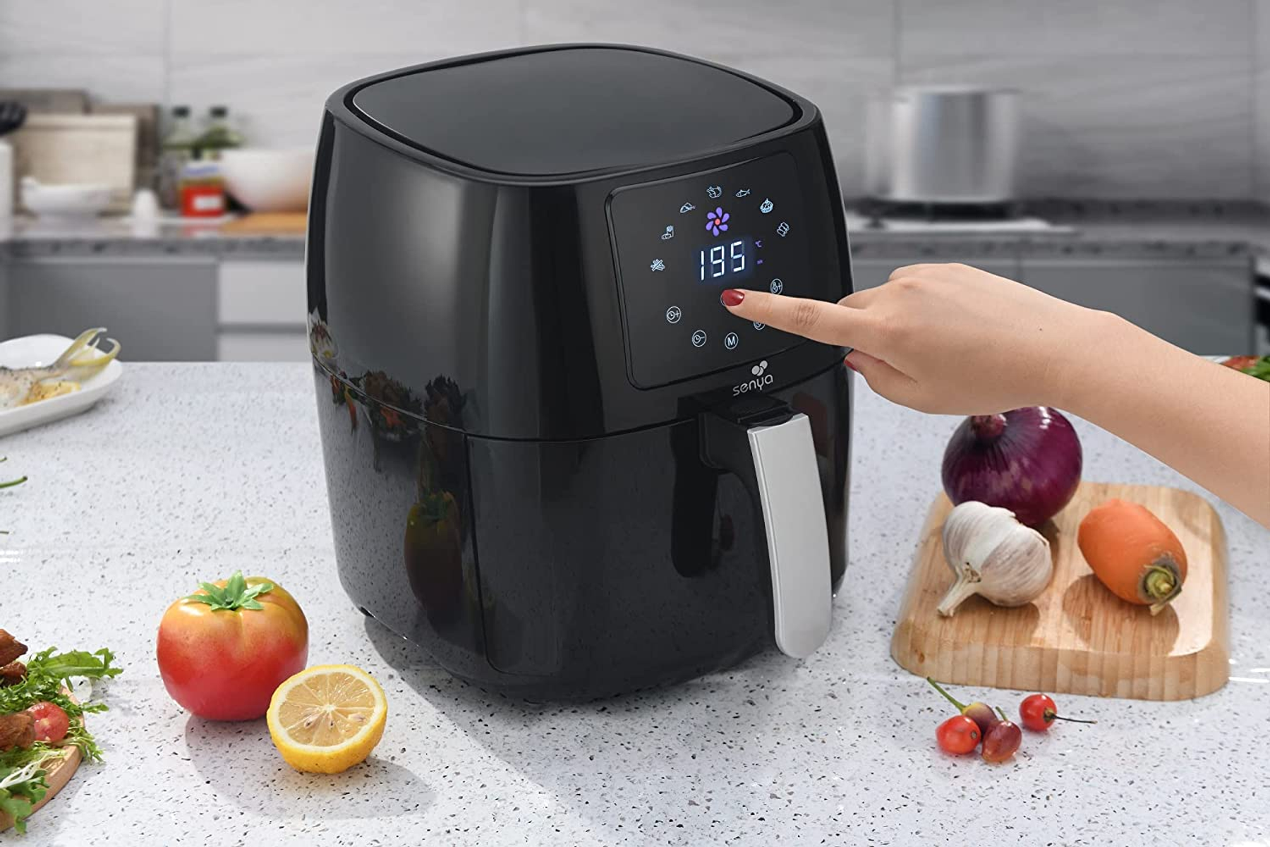 Friggitrice ad aria nera con mano che tocca il display digitale. Verdure e frutta intorno.