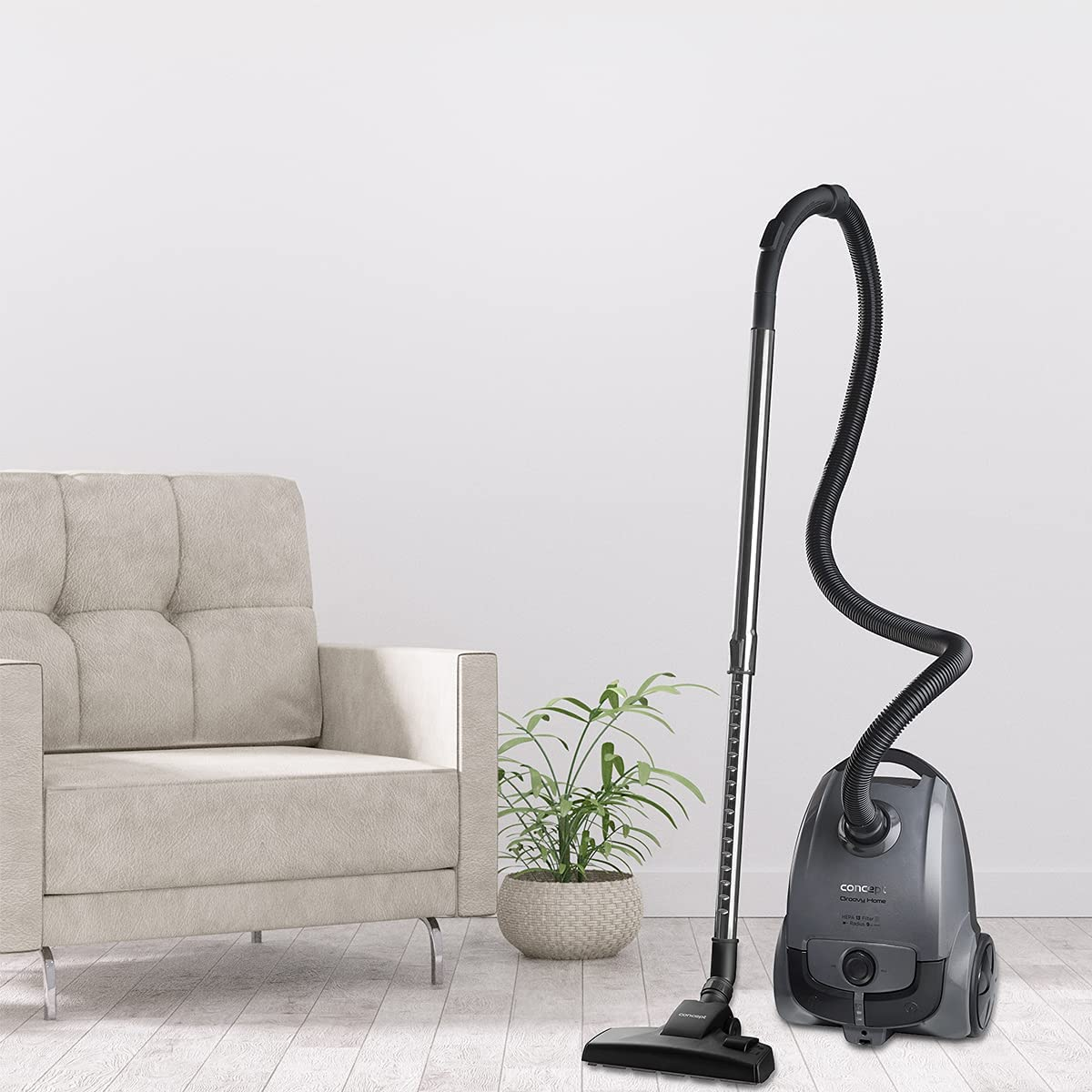 A gray vacuum cleaner standing upright next to a plant, armchair and a wall.