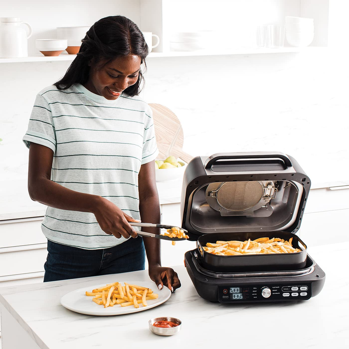 Donna usa le pinze per mettere le patatine dalla friggitrice su un piatto. Ambiente cucina, patatine con ketchup.