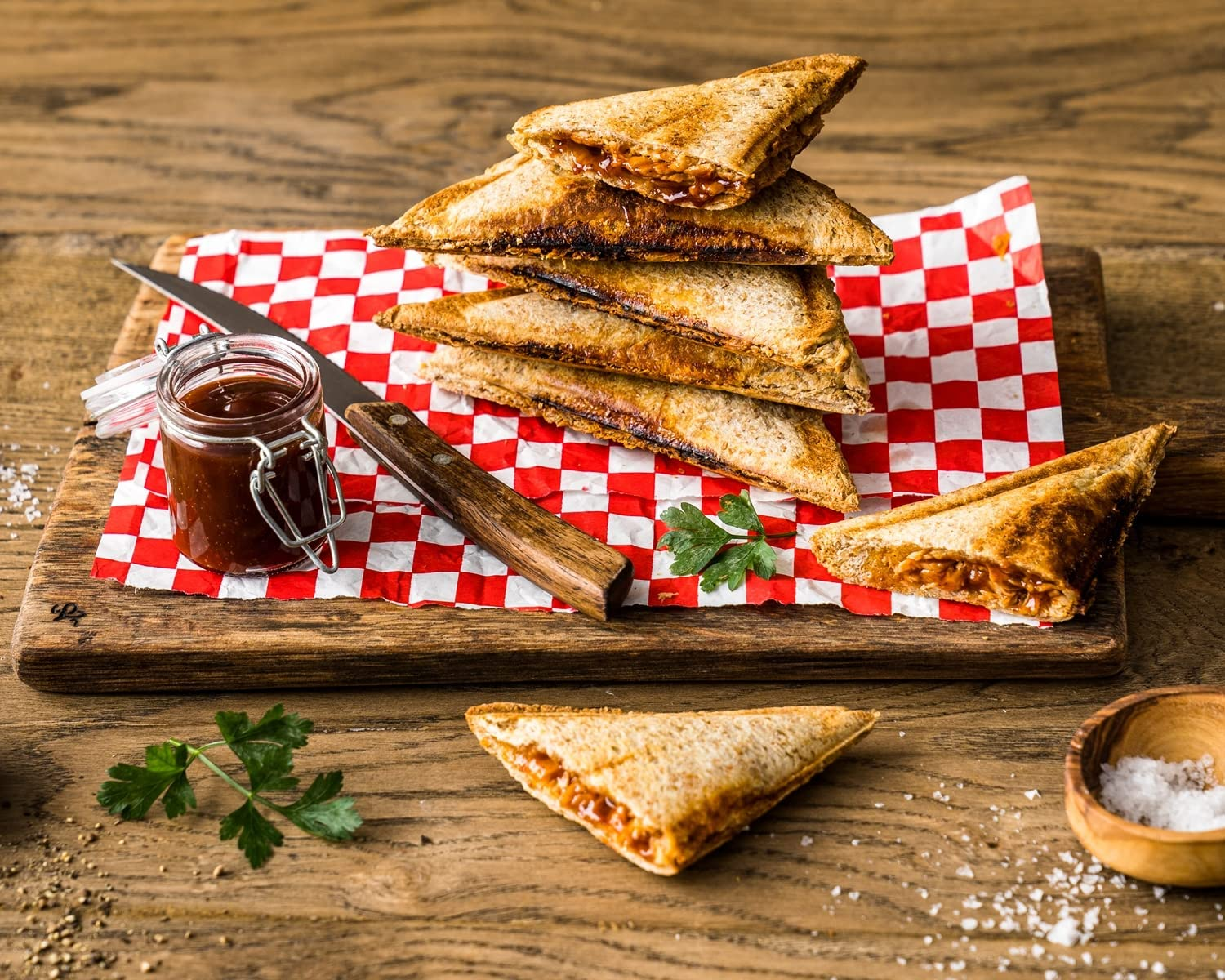 Stos tostowanych kanapek na czerwono-białej kratce, z nożem i słoikiem sosu.