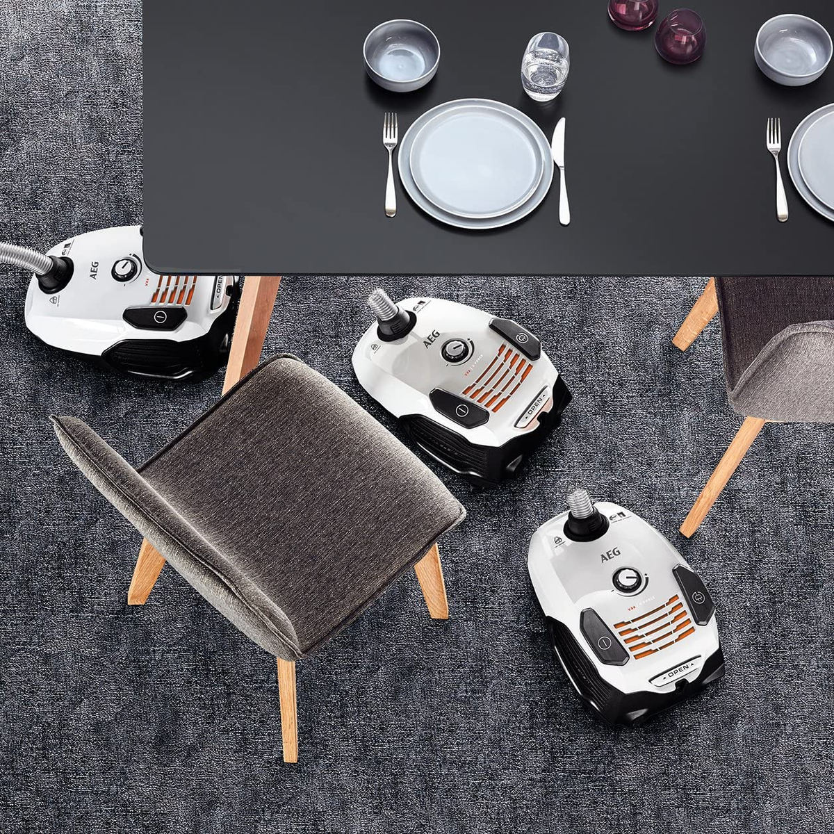 Nakrycie stołu: talerze, sztućce i szklanki. Odkurzacze i krzesła ustawione wokół czarnego stołu na szarej wykładzinie.